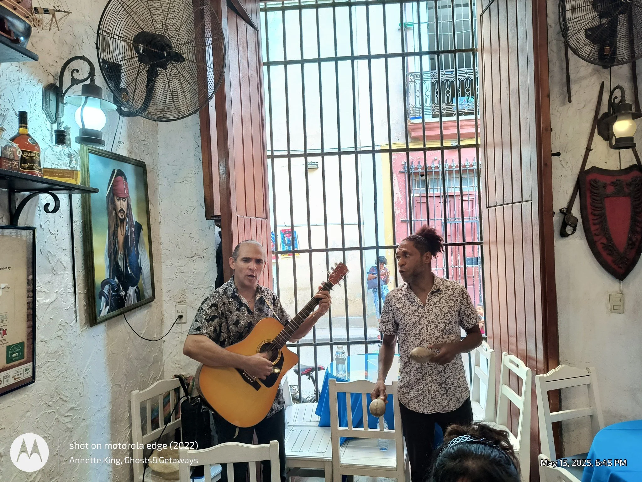 Two men performing music indoors, one playing the guitar and the other holding percussion instruments, with a barred window and various wall decorations behind them.