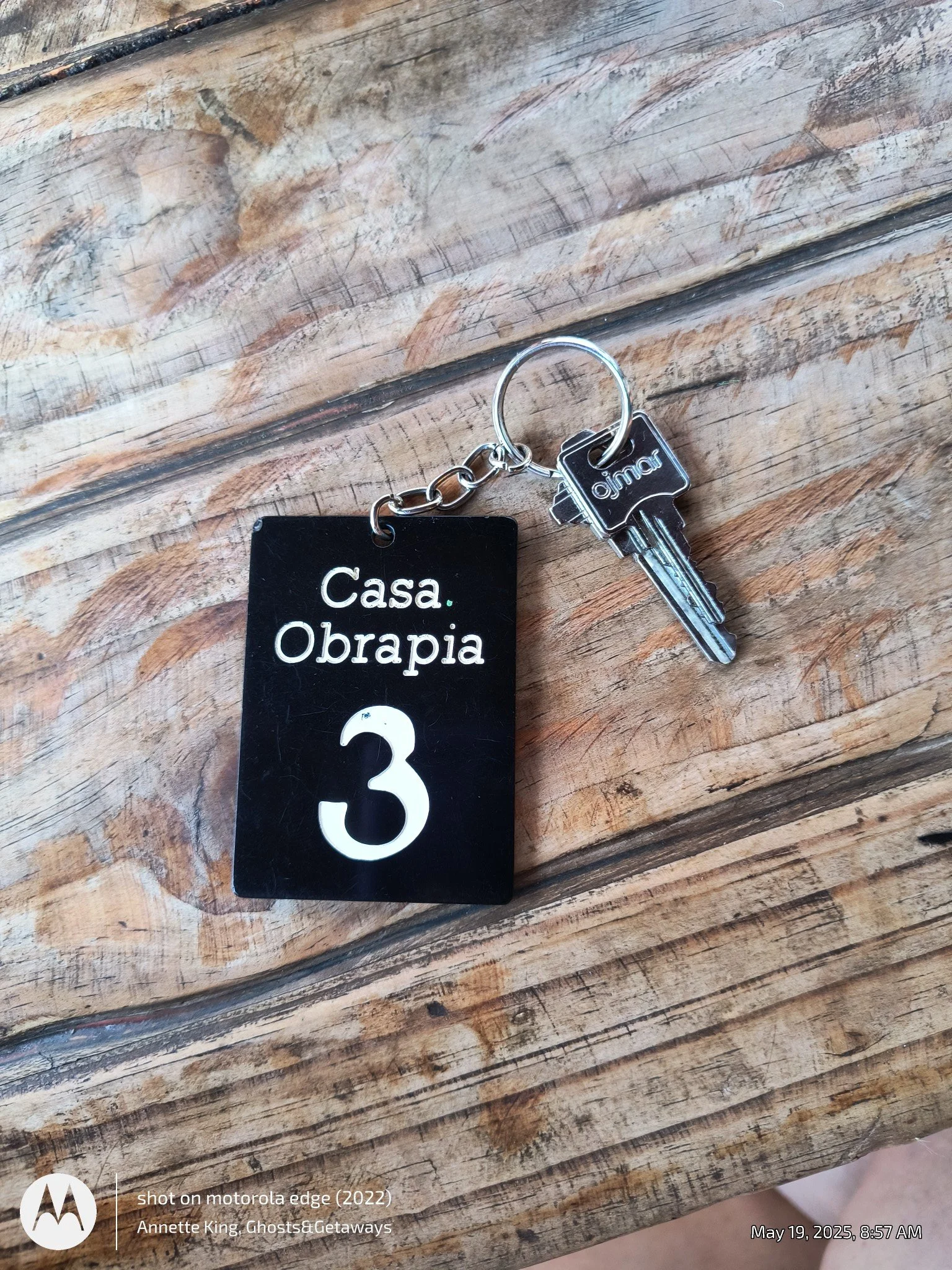 A set of keys attached to a black tag with white text that says 'Casa. Obrapia'. The keys include a car key with a black plastic head branded 'aimo' and a silver metal key. The background is a wooden surface with visible grain and cracks.