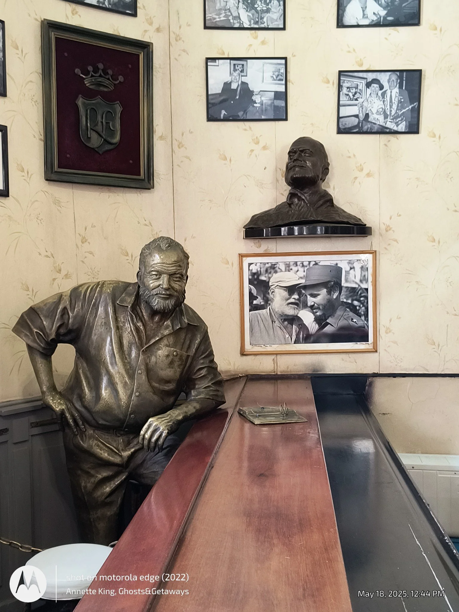 Bronze bust of a historical man on a wooden table with framed black-and-white photographs and artwork on the wall behind.
