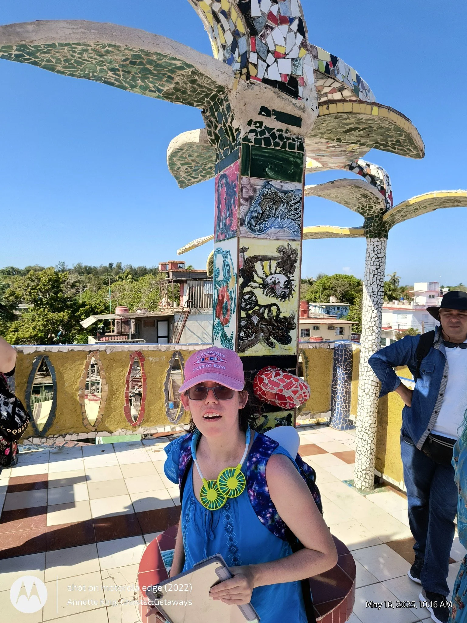 A woman with a blue shirt, sunglasses, and a pink cap holding a smartphone, standing in front of a colorful mosaic sculpture with the sky and trees in the background. Several other people are partially visible around her.