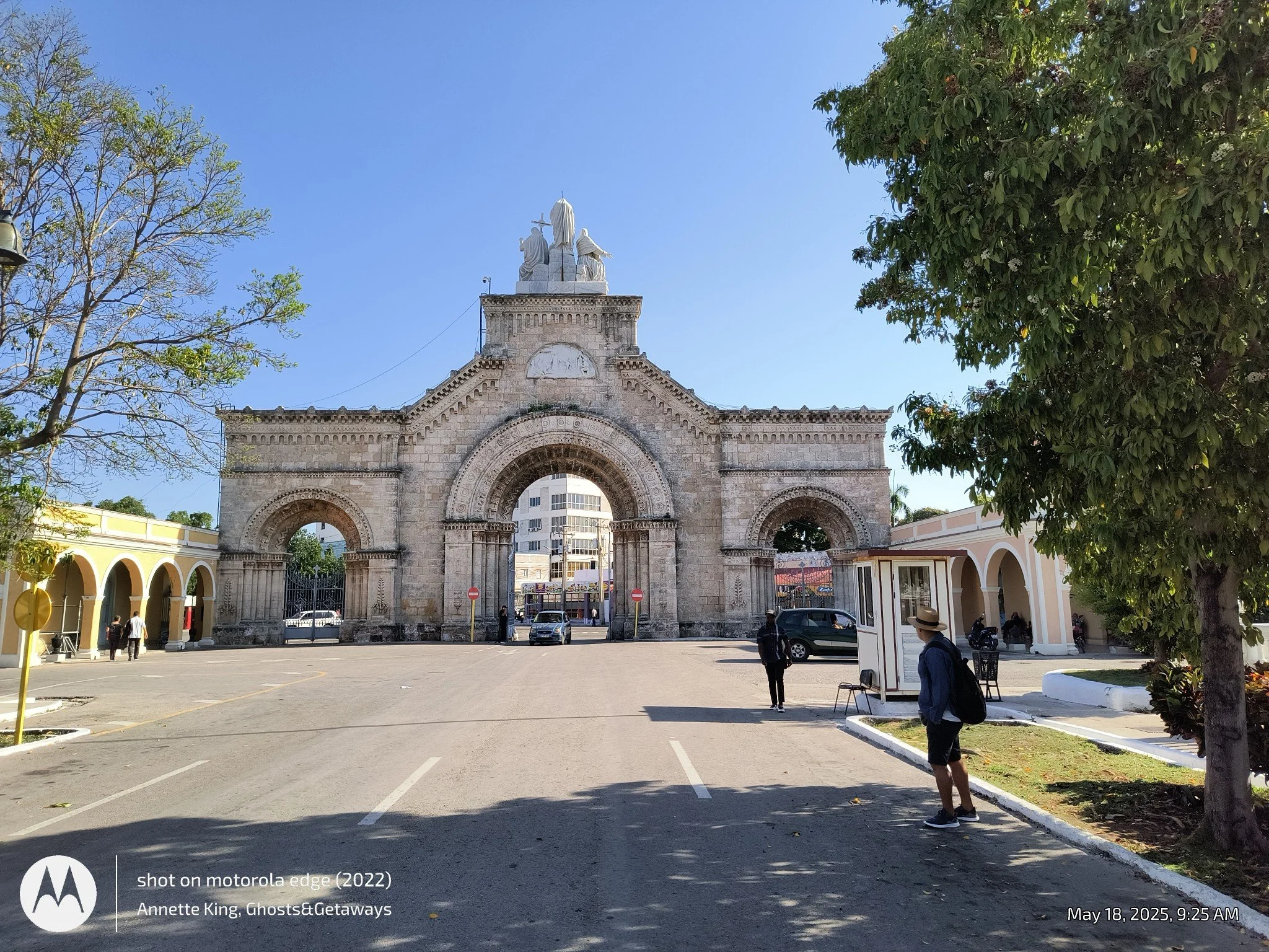 View of an arched stone monument with three statues on top, set in a plaza with trees, pedestrians, and a bright blue sky.