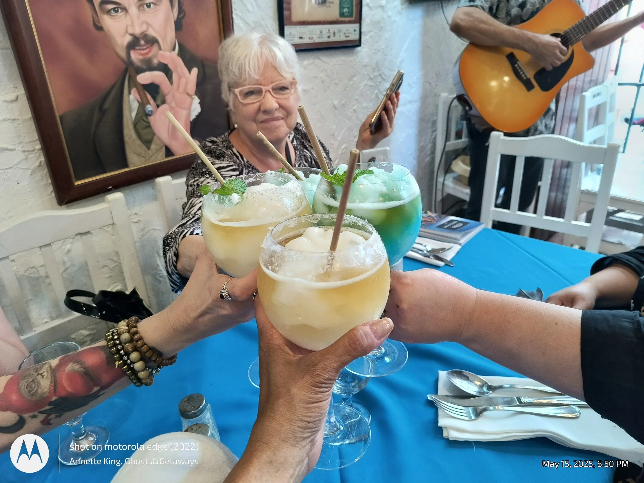 Four people are celebrating with colorful cocktails in a restaurant. The drinks are garnished with mint leaves and have straws. A woman with white hair, glasses, and a patterned top is smiling, and another person with a tattooed arm is holding a drin