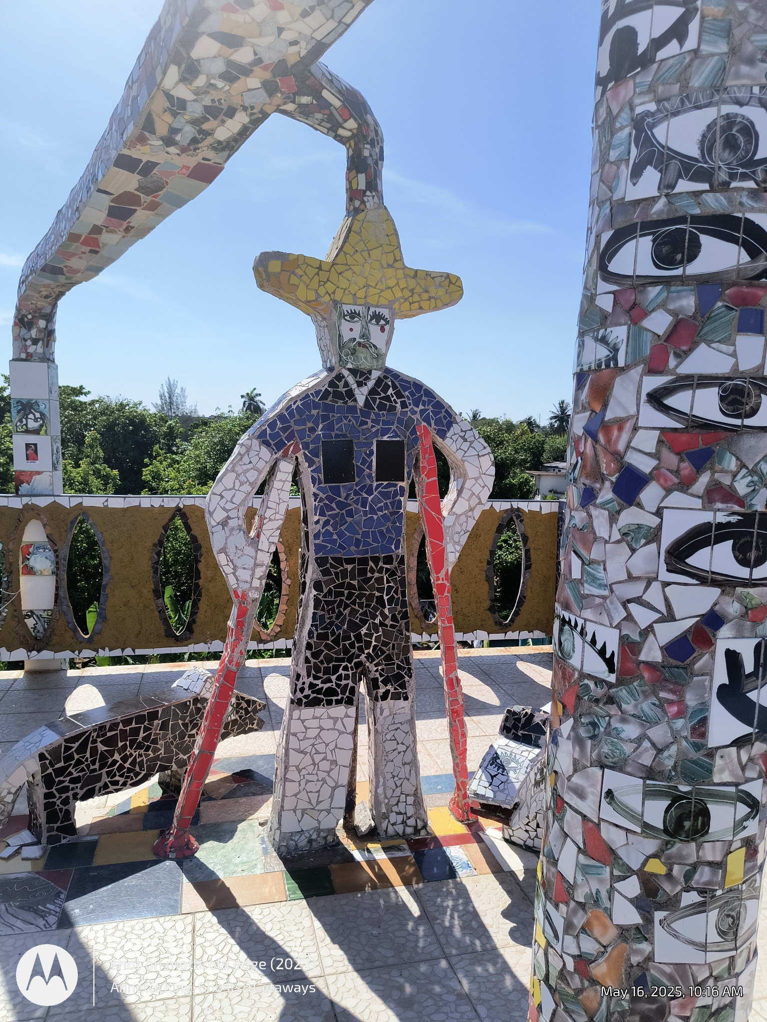 A mosaic sculpture of a man with a yellow sombrero and a mustache, standing with one hand on his hip in an outdoor park, with trees and a blue sky in the background.