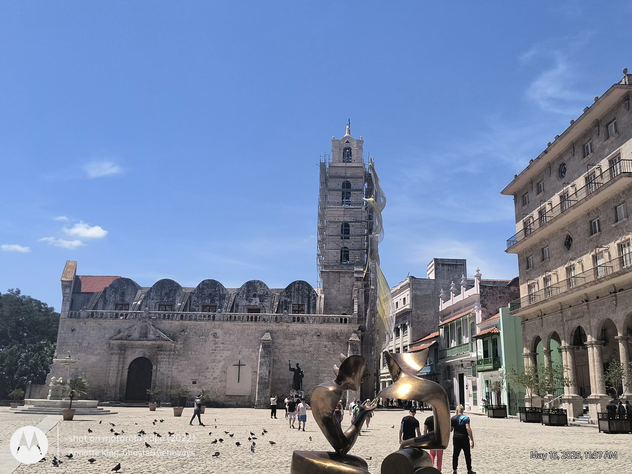 A historic church with a tall bell tower under renovation, with scaffolding and yellow protective sheeting. In front, there are modern buildings, a metallic sculpture of abstract art, and people walking in a square with pigeons on the ground. The sky
