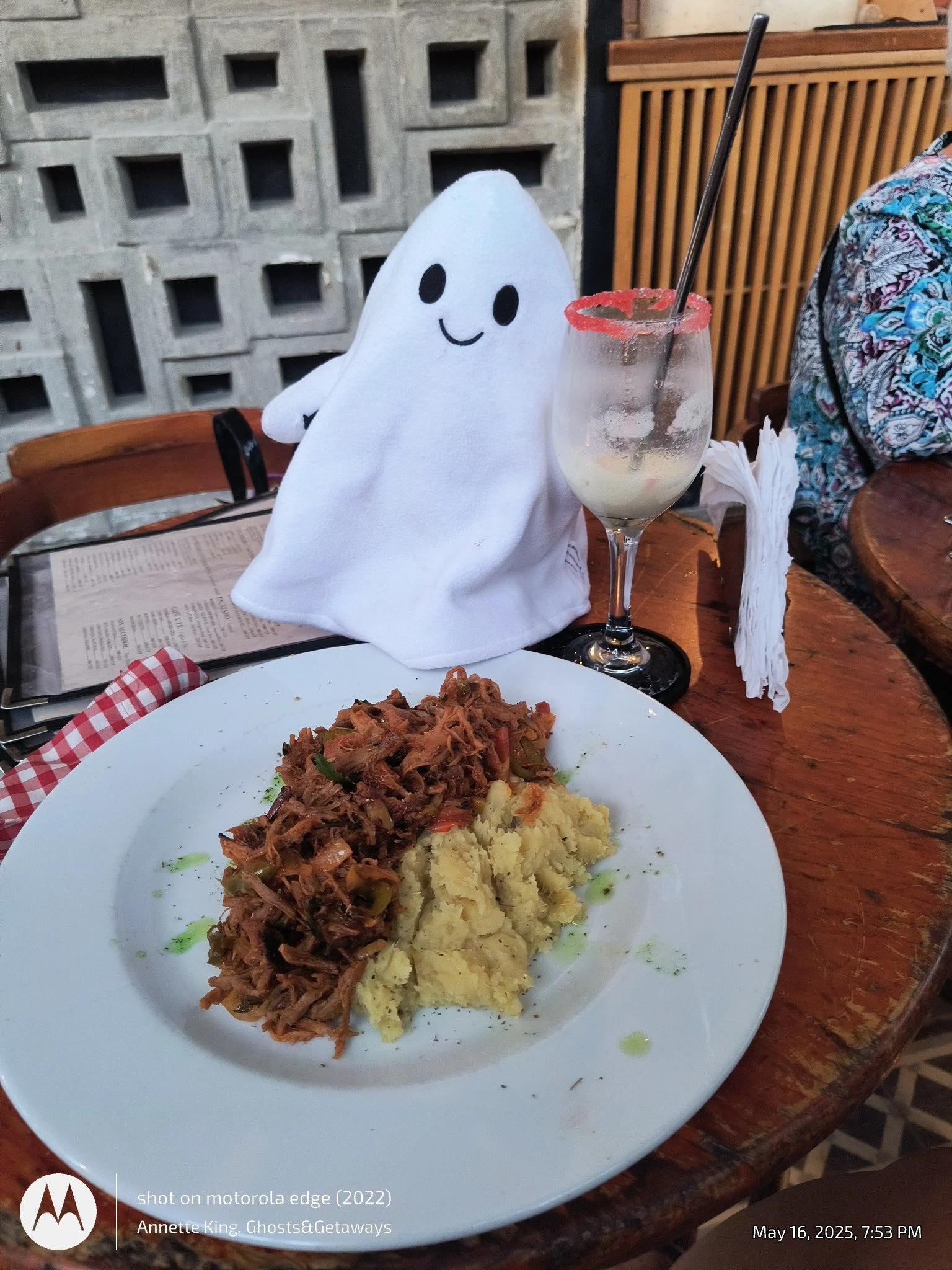 A plate of mashed potatoes with shredded beef and vegetables on top, served on a white dish. In the background, there is a glass of a creamy beverage with a straw, a doll in the shape of a ghost with a smiling face, and a wooden table with a menu and