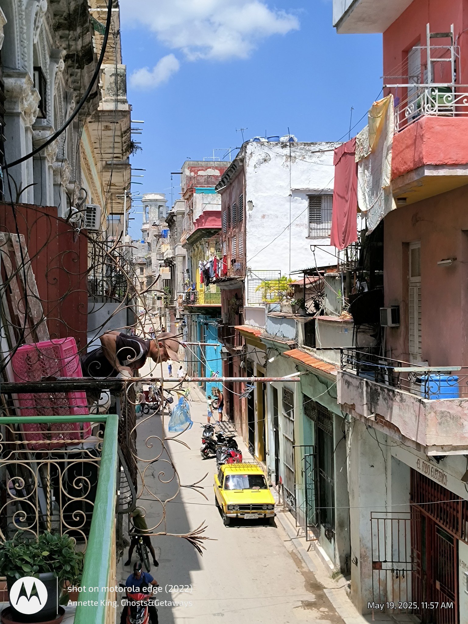 A narrow street in a residential area with colorful, worn buildings on both sides. Clothes are hanging on lines outside the apartments. Several parked motorcycles and a yellow taxi are visible on the street. A person is seen leaning over a balcony ra