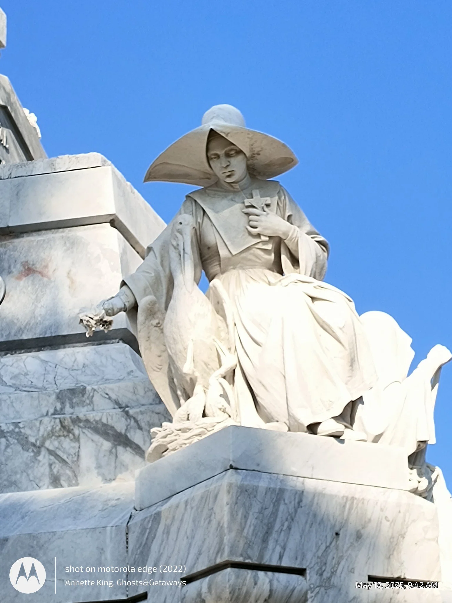Close-up of the marble sculpture of a seated woman with a large hat, holding a cross and a book, against a clear blue sky.