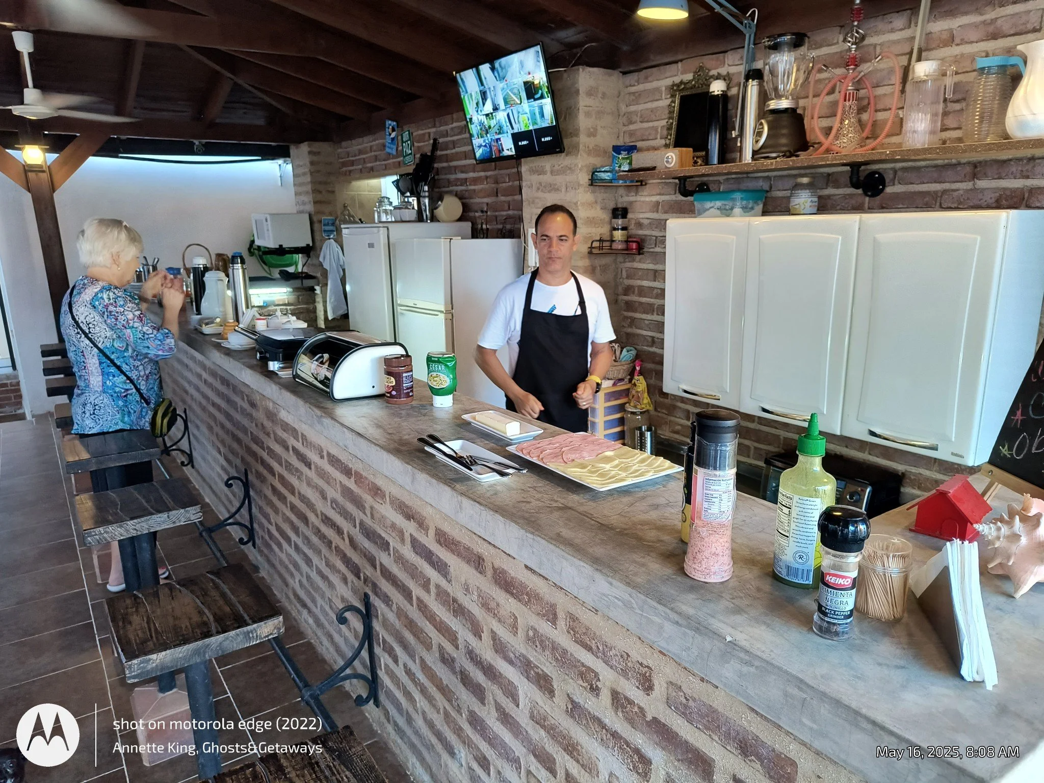 A restaurant kitchen with a brick wall and a counter where a man wearing an apron is preparing food, with various ingredients and supplies. An elderly woman is sitting at the counter eating, and a flat-screen TV is mounted on the brick wall.
