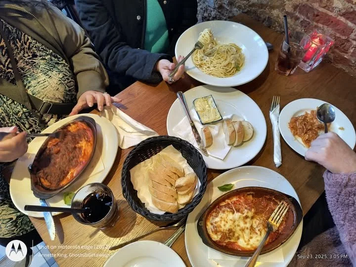 A top-down view of a wooden table with various dishes including two baked pasta dishes, sliced bread, a small dish of butter, a plate with a slice of bread, a glass of red wine, and some drinks, surrounded by four people reaching for the food.