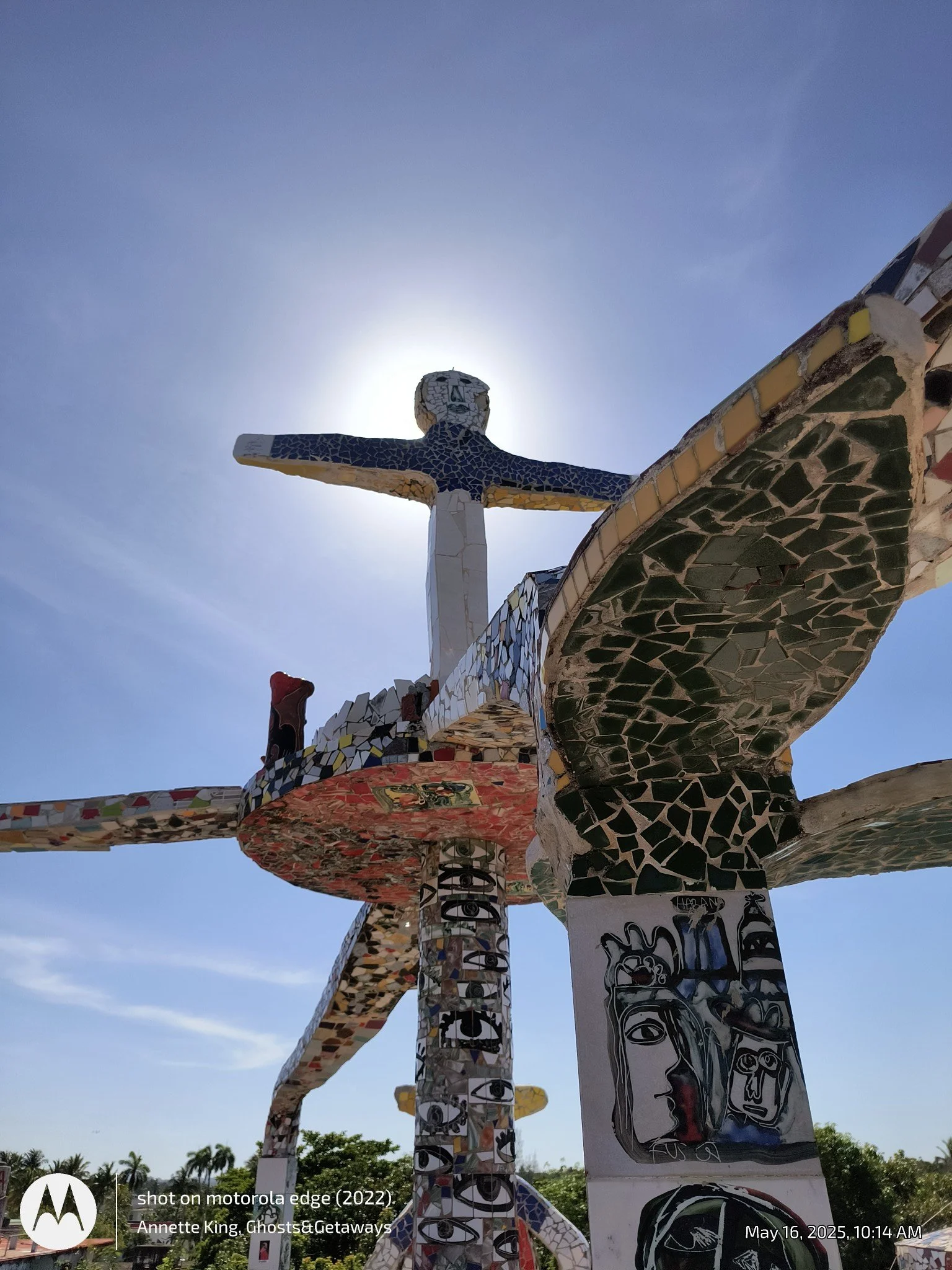 Mosaic sculpture of Jesus on a cross with arms outstretched, set outdoors with blue sky and trees in the background.