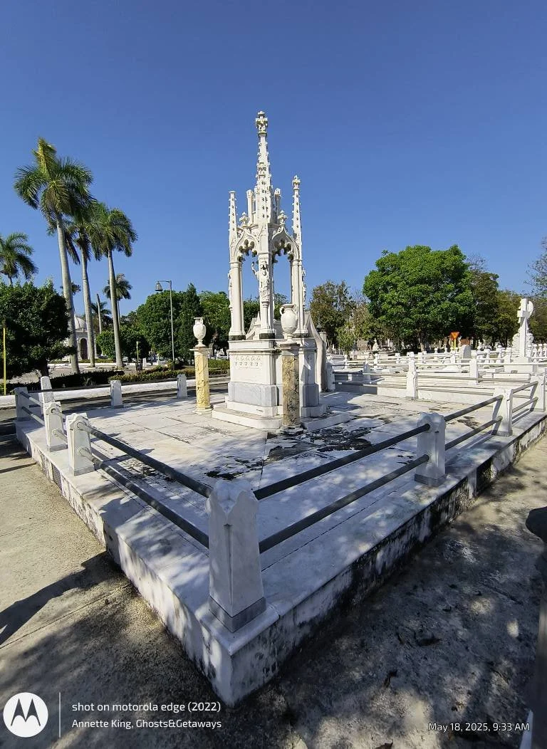 A white marble monument with intricate gothic architecture, surrounded by a low fence, in a park with palm trees and green trees under a clear blue sky.