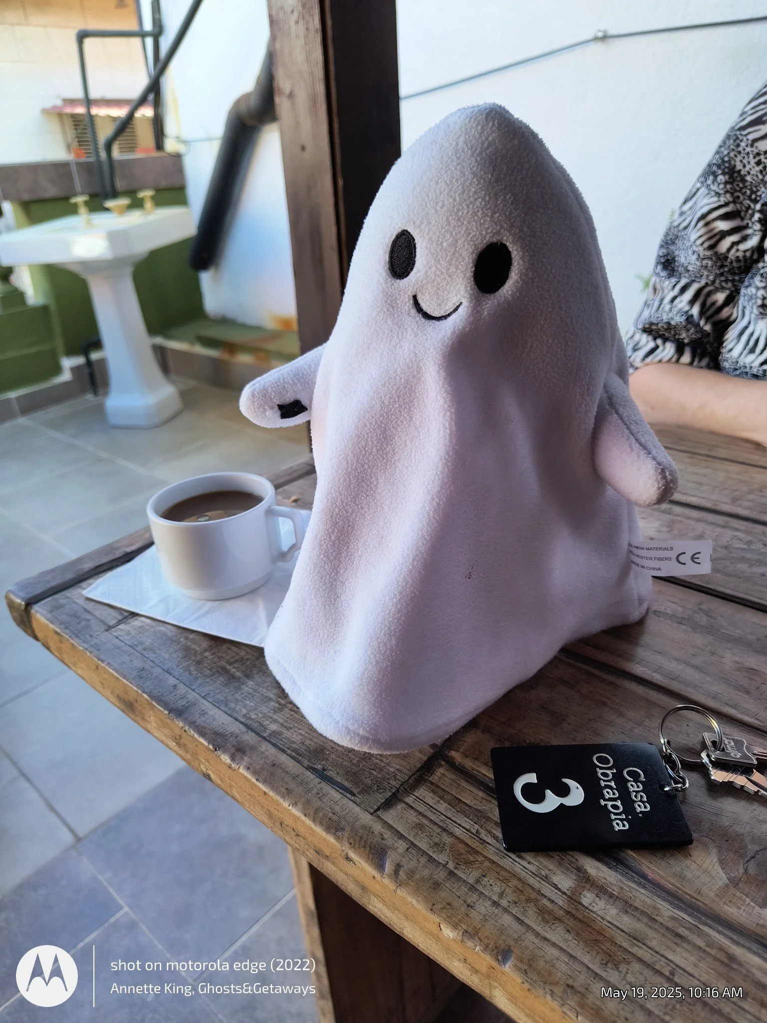 A small plush toy with a white, ghost-like shape and black facial features, perched on a wooden table next to a white mug of coffee. The background includes a white wall, a window, and a person wearing a patterned shirt. There is also a black keycard