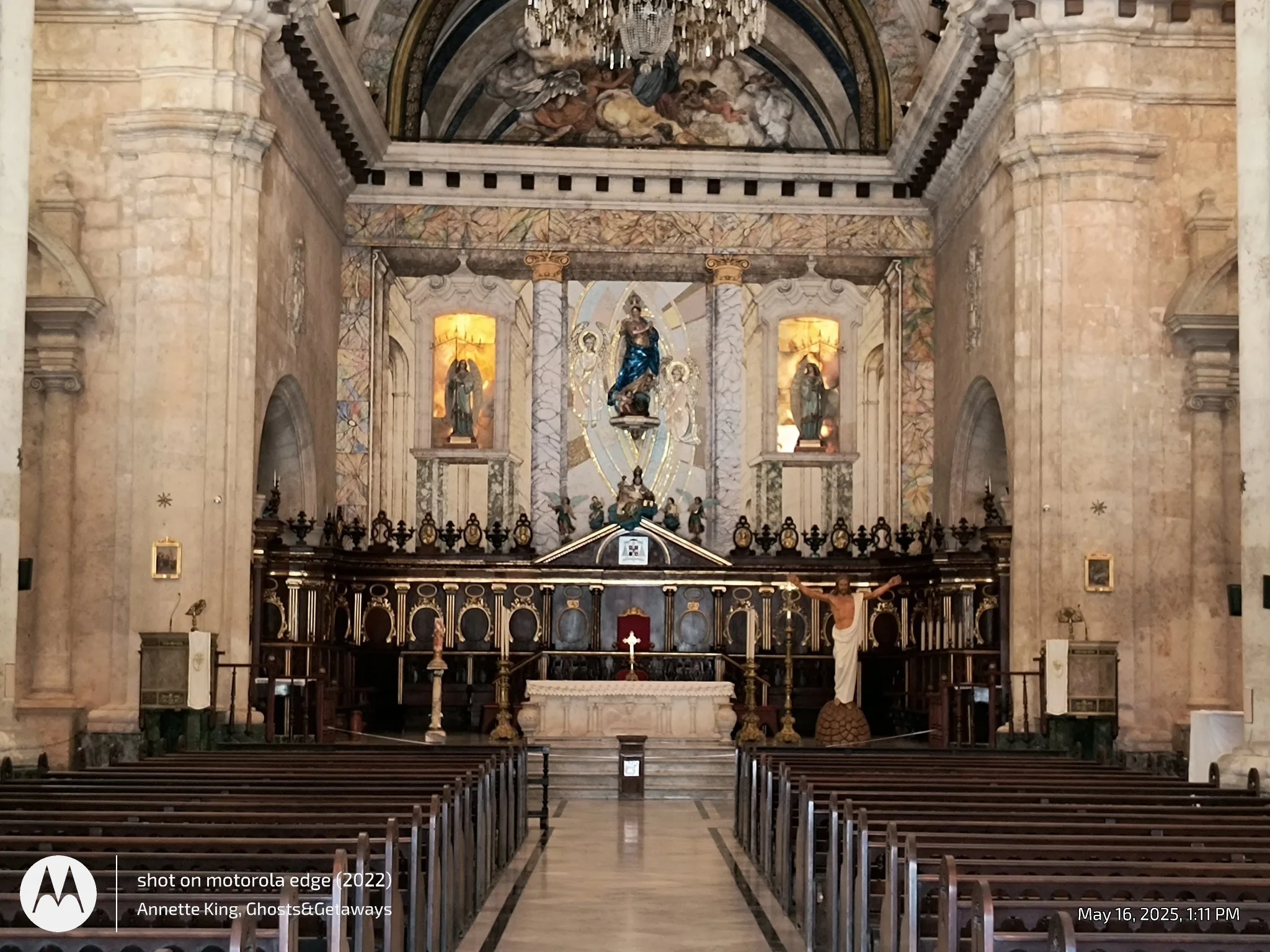 Interior of a church or cathedral with wooden pews, an altar with religious statues and a crucifix, marble columns, and ornate ceiling artwork including a fresco painting.