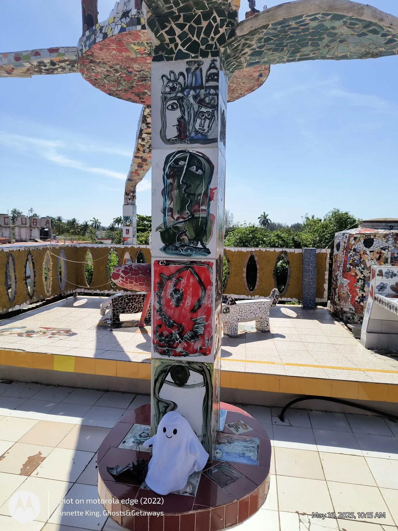 Colorful mosaic sculpture with various framed abstract art pieces and a plush ghost figure on the base. The sculpture is situated outdoors under a clear sky with trees and a yellow railing in the background.