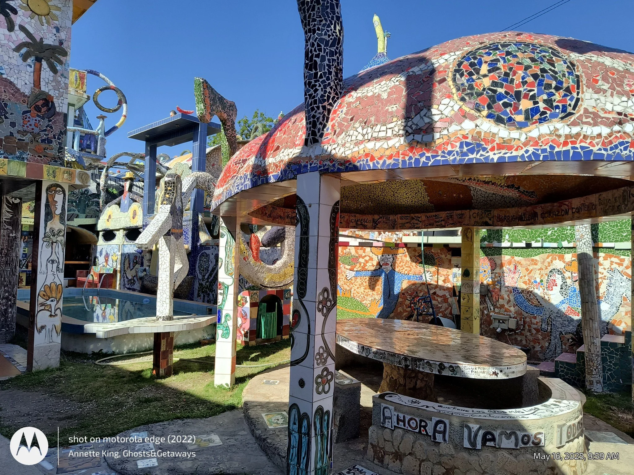 Colorful mosaic sculptures and structures in a park, including a large serpent head and various whimsical shapes, with bright sunlight and a clear blue sky.