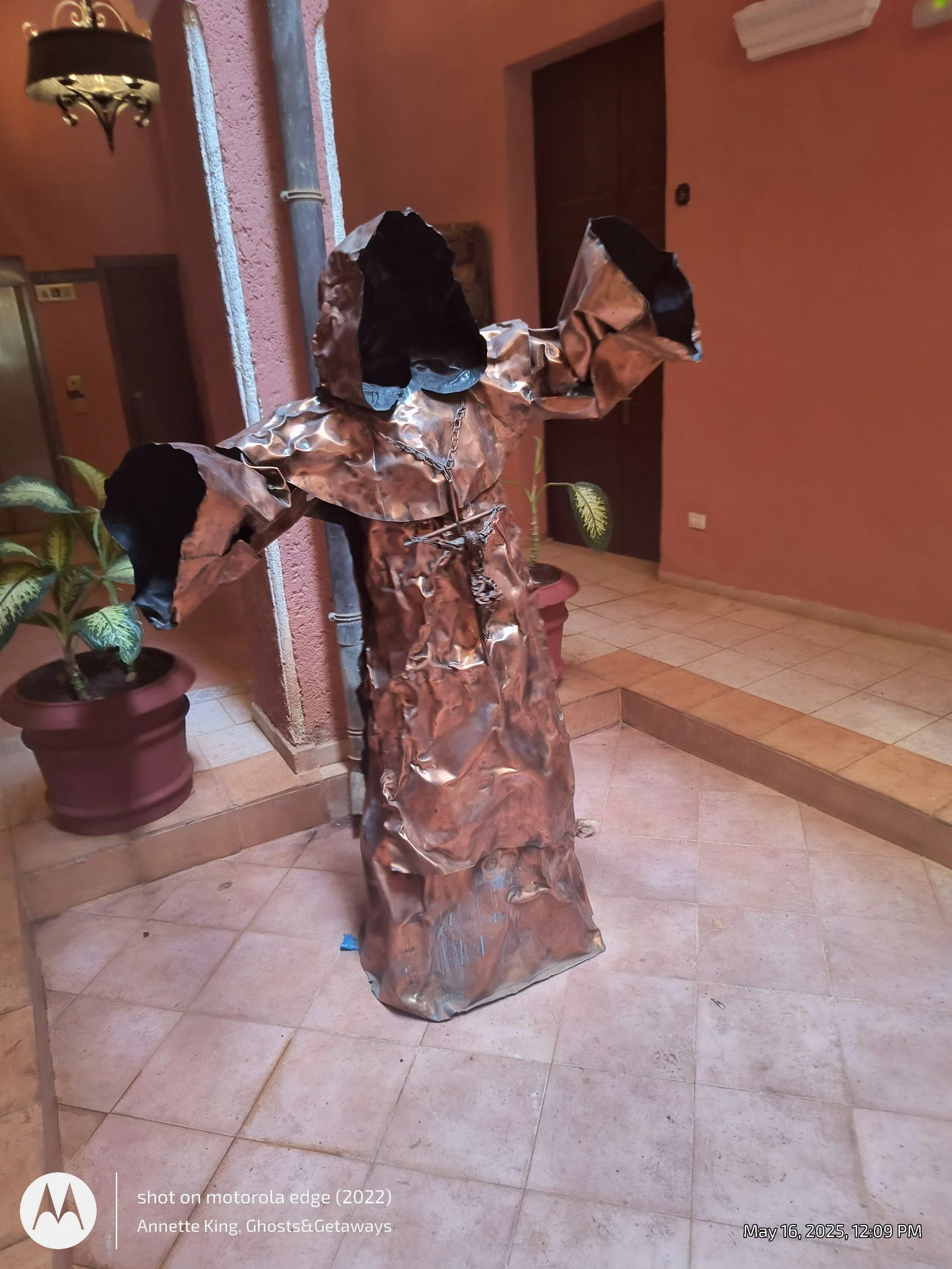 A humanoid sculpture made entirely out of crumpled, shiny metallic material standing in an indoor setting with pink textured walls. The sculpture has a chain around its neck and black fabric or material covering its head. Potted plants and interior f