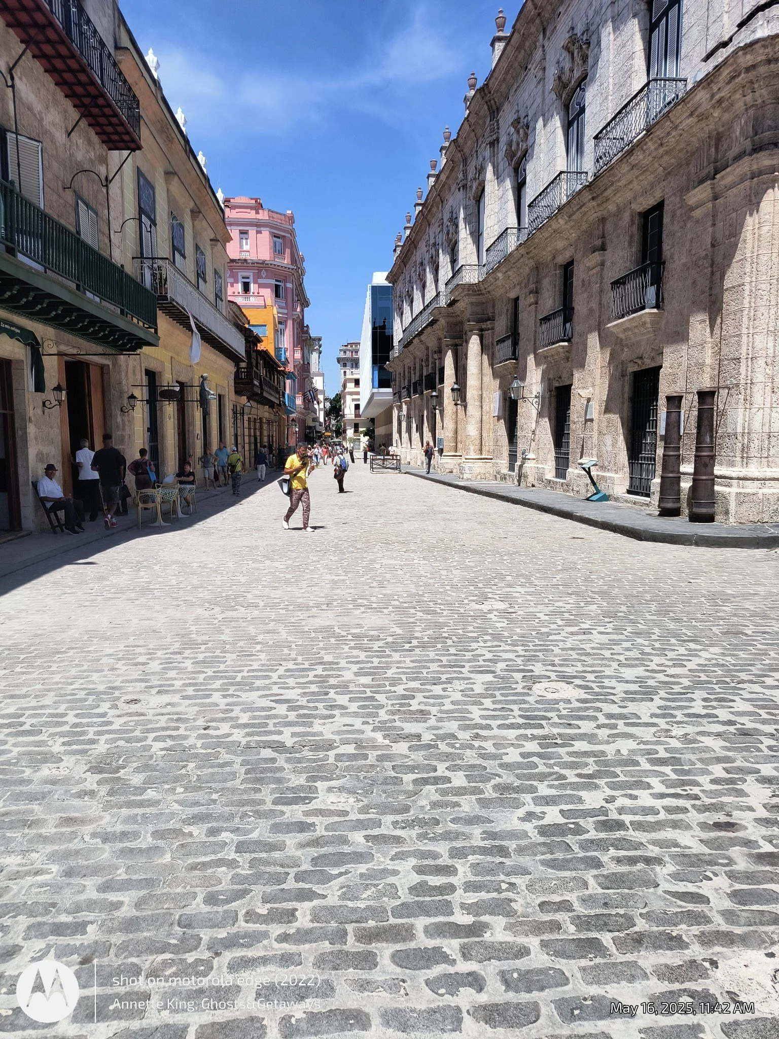 A street scene with cobblestone pavement, modern and historic buildings on both sides, and people walking and sitting at outdoor tables. The sky is clear and blue.