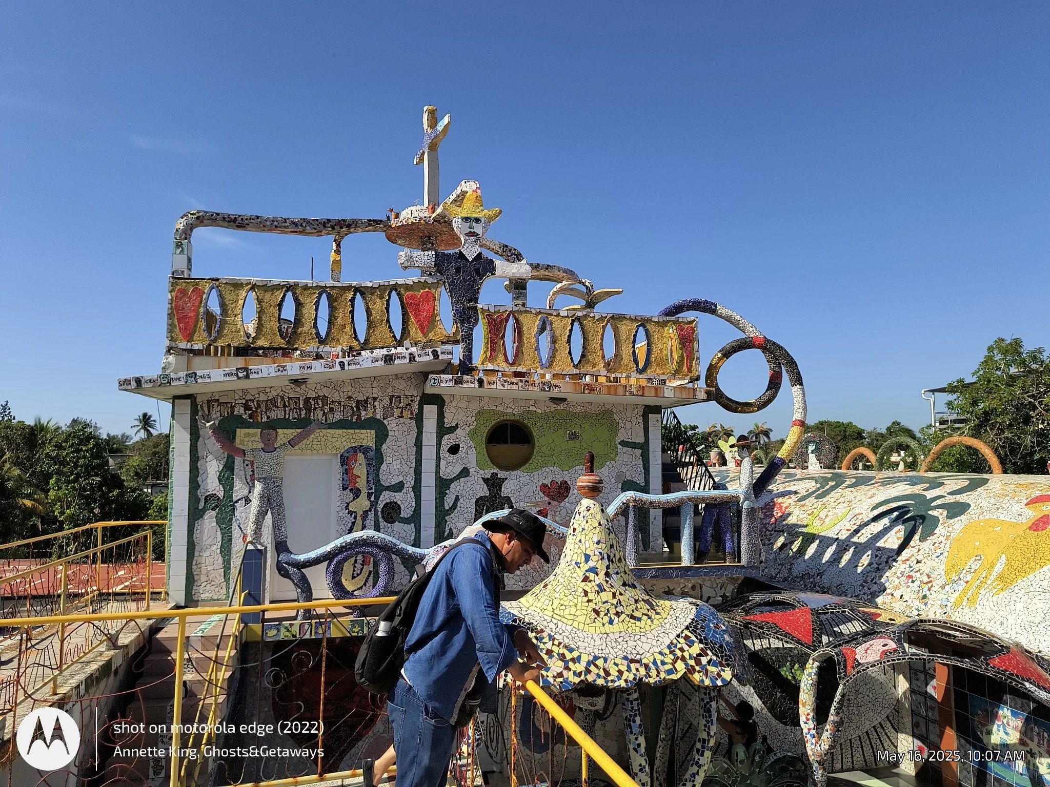 A colorful mosaic sculpture of a person riding a bicycle, with a woman wearing a wide-brimmed yellow hat and a man with a backpack looking at the sculpture. The sculpture has a woman with blonde hair, detailed facial features, and is decorated with m