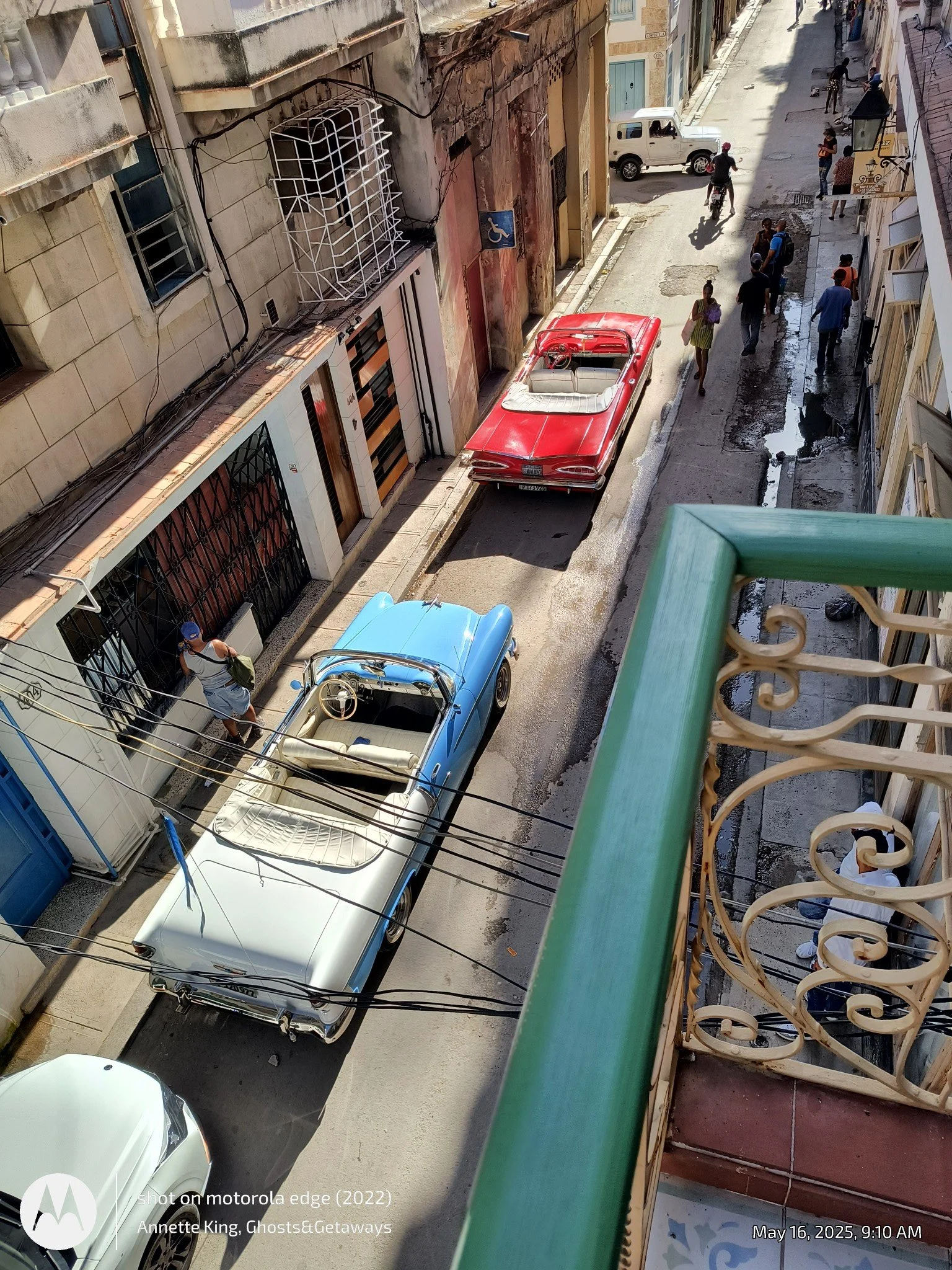 A view of a narrow urban street from a balcony, showing a vintage red convertible car parked on the side, several pedestrians walking, a white SUV, a motorbike, and buildings with worn facades. The street appears slightly wet or patched in some areas