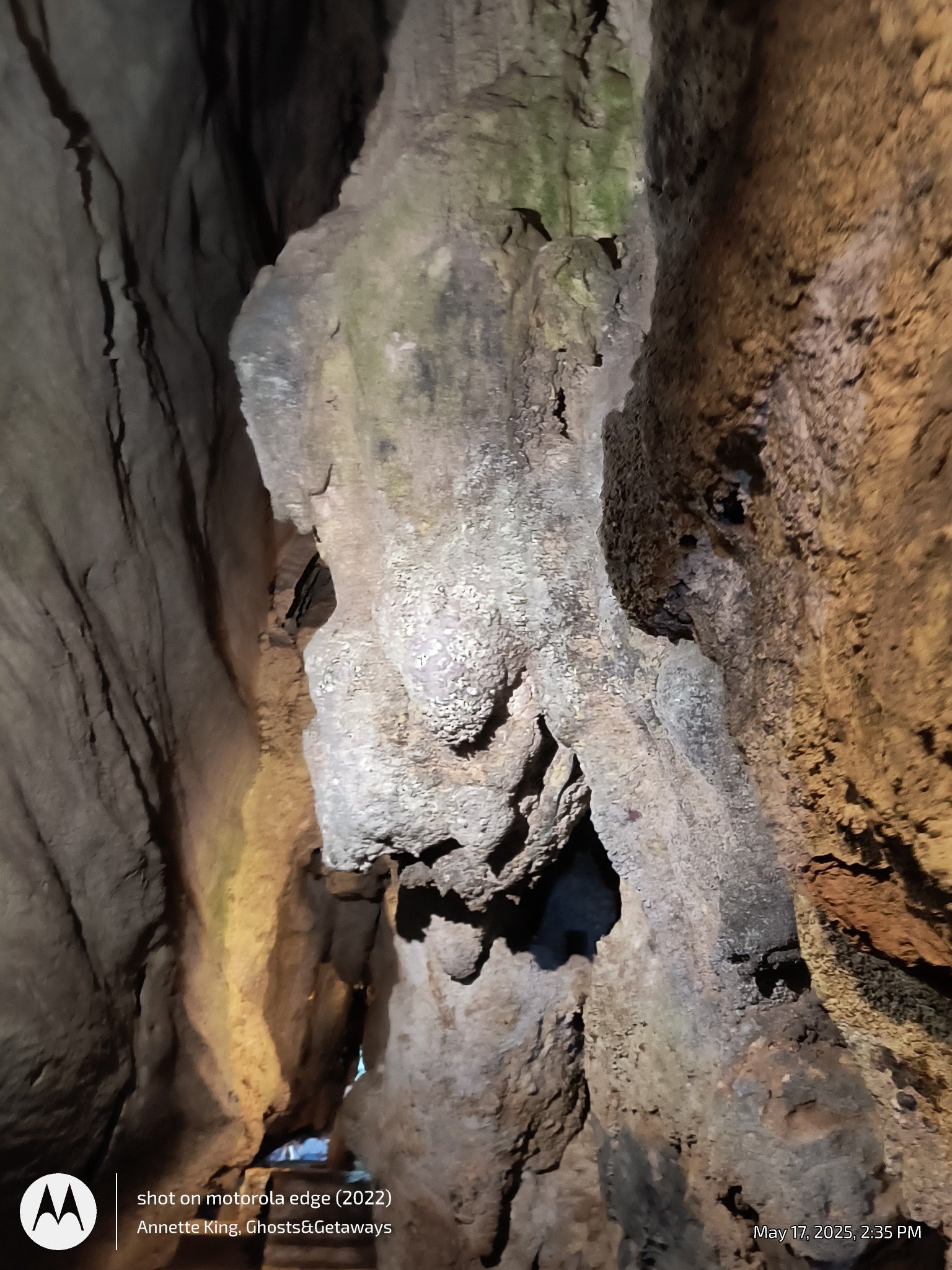 Inside a cave with rugged, uneven stone walls and a dirt floor. There are openings in the cave walls allowing some natural light to enter.