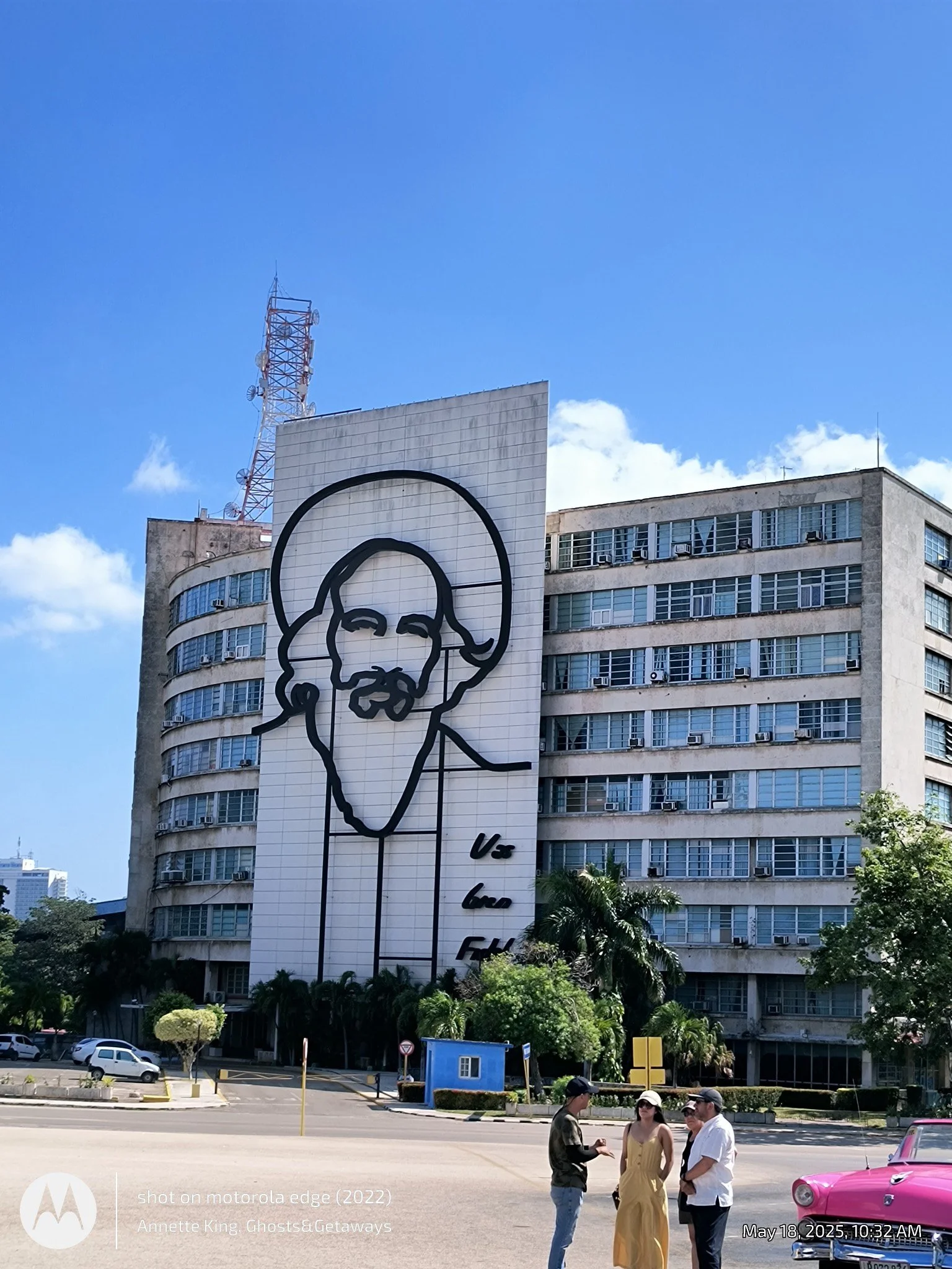 A tall building with a billboard of a drawn face, and a group of four people standing and talking in the foreground, with classic cars parked nearby, under a clear blue sky.