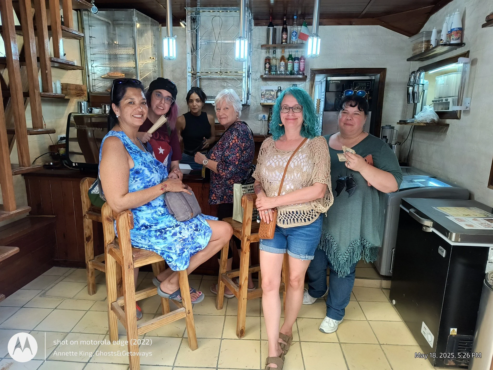Six women and one young girl standing and sitting in a restaurant kitchen area, smiling and posing for the photo. The woman seated in the foreground wears a blue floral dress, glasses, and carries a bag on her lap. The woman standing to her right has