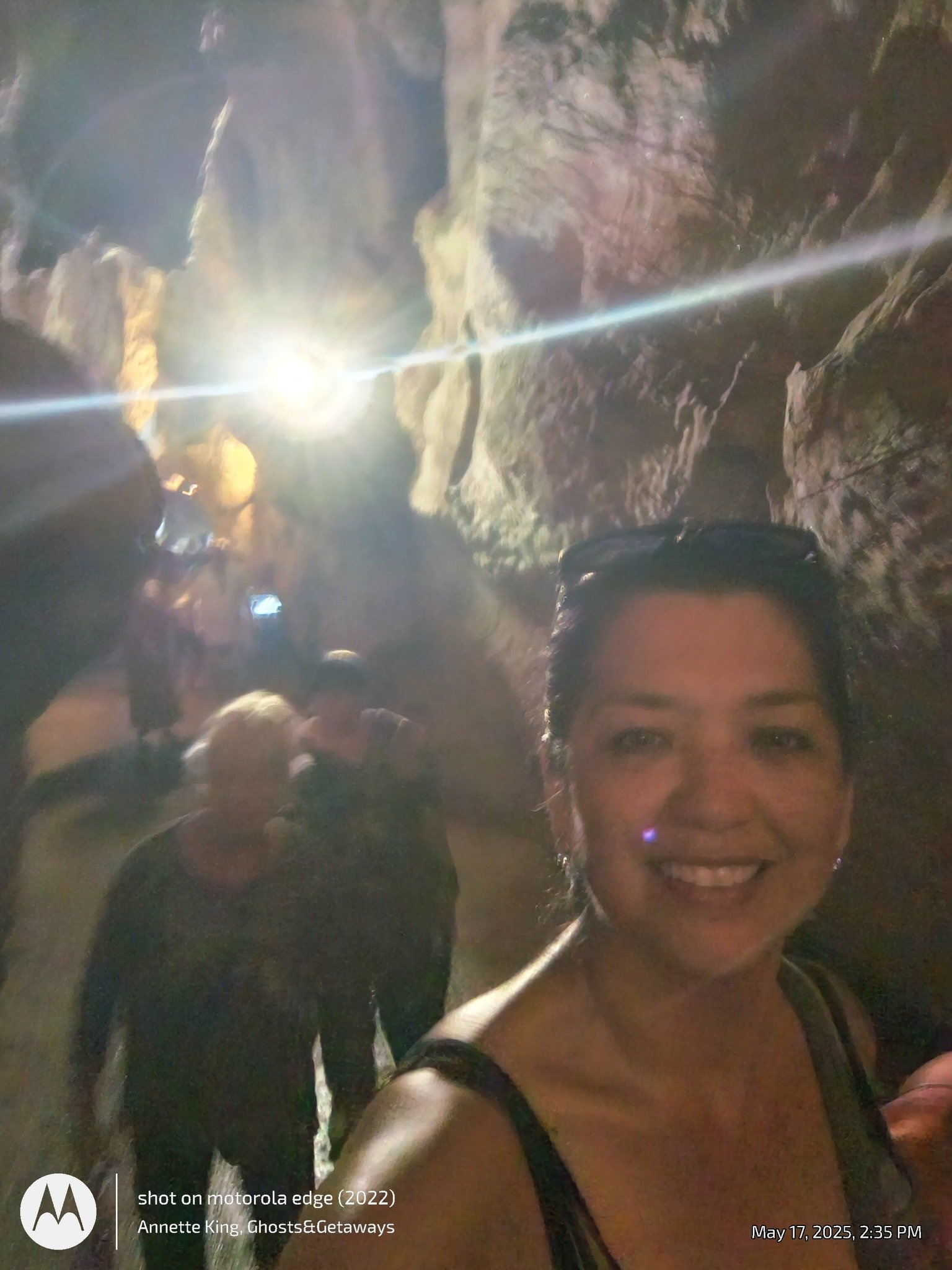A smiling woman taking a selfie in a cave or underground cavern with a waterfall flowing behind her.