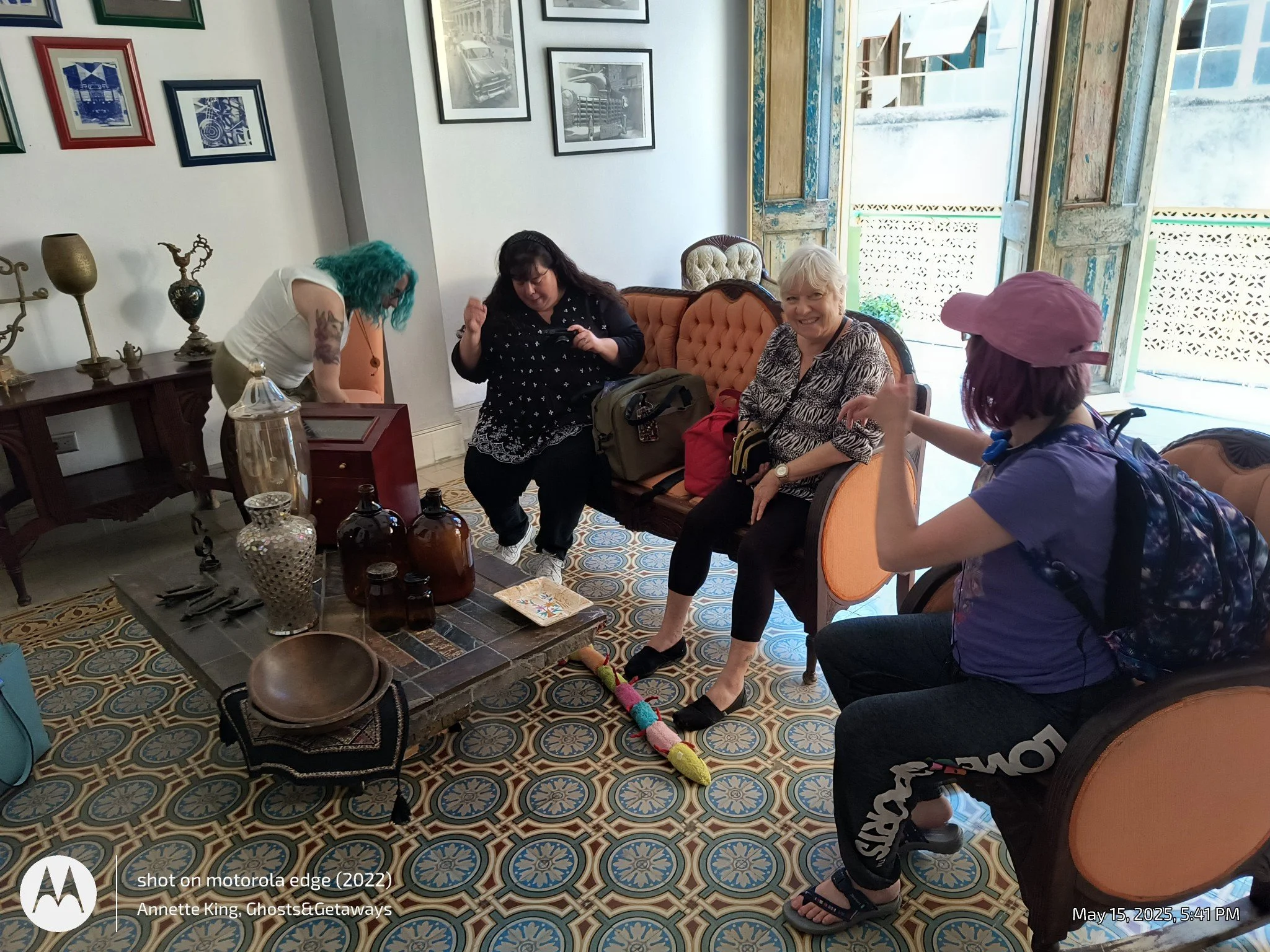 Four women sitting in a living room with vintage decor, some wearing hats, with a table in front holding bottles and decorative items, and a large open door leading outside.