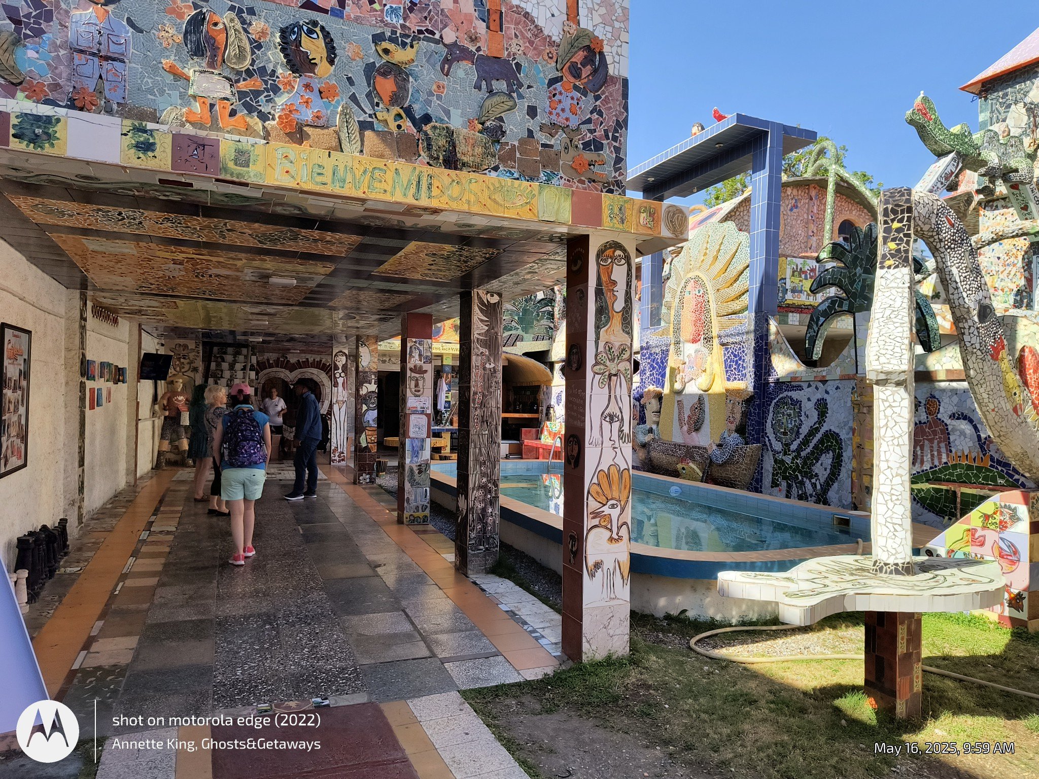 Colorful outdoor mosaic art installations featuring various animal and human figures, with bright blue sky in the background and several people walking underneath a decorative archway.