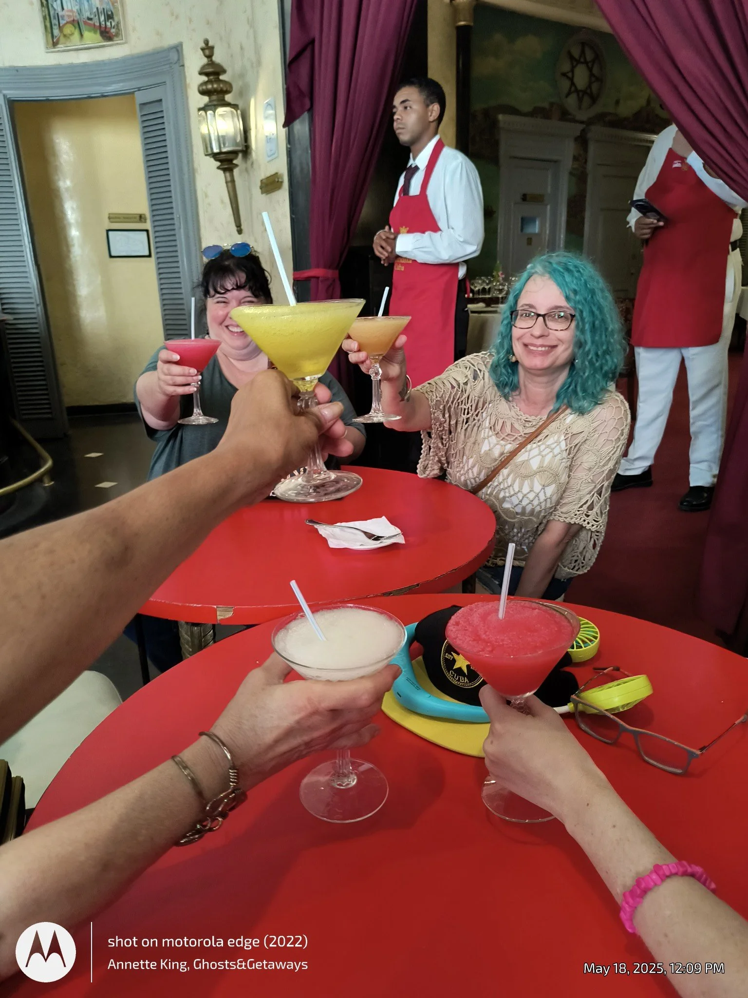 People sitting at a red table enjoying drinks, with hands holding various cocktails in the foreground. Two women with glasses and colorful hair are smiling, and waitstaff in red aprons are visible in the background inside a restaurant.