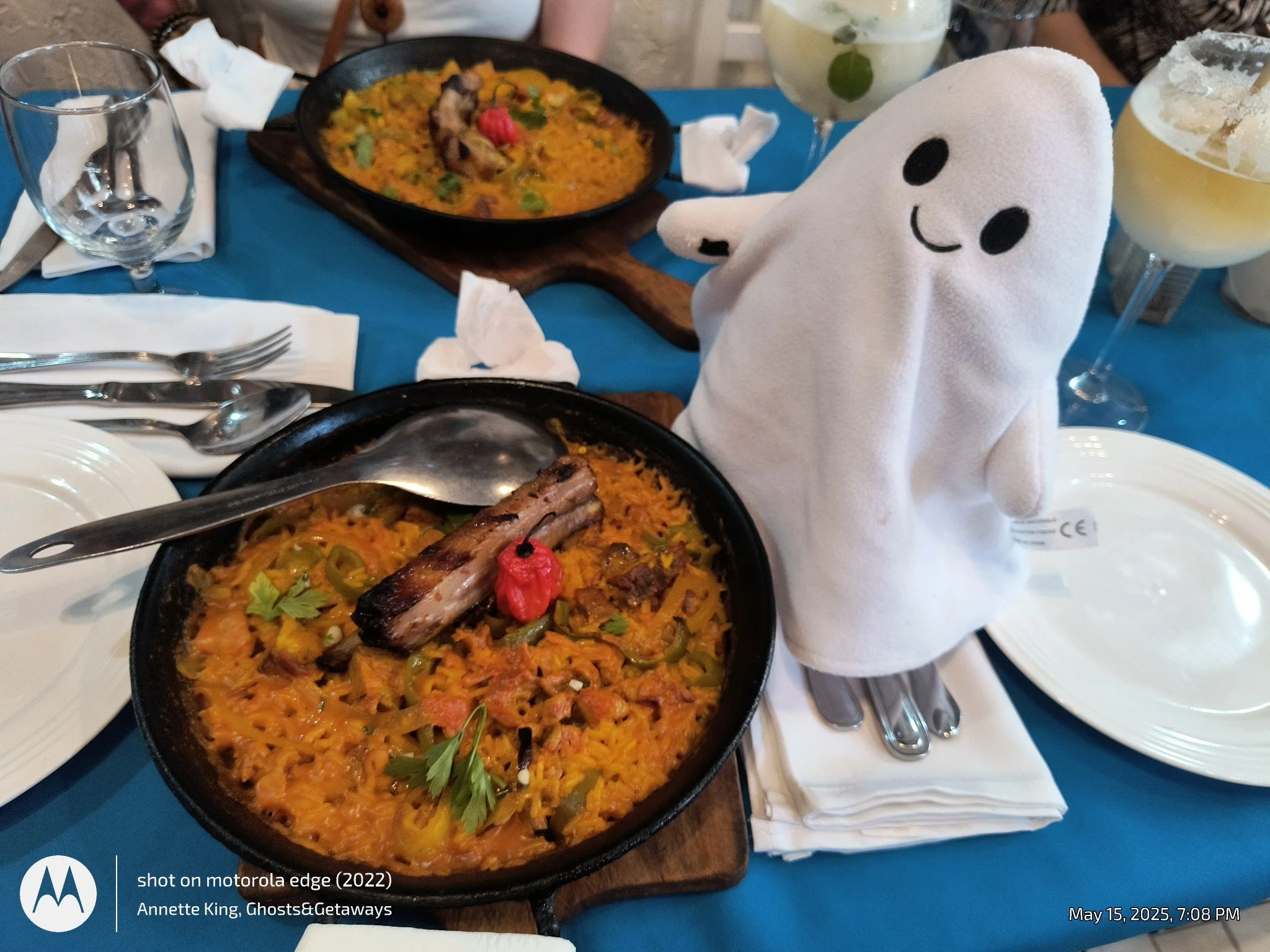A table set with two bowls of yellow rice with vegetables and grilled meat, surrounded by plates, utensils, and glasses, with a plush ghost toy on the side.