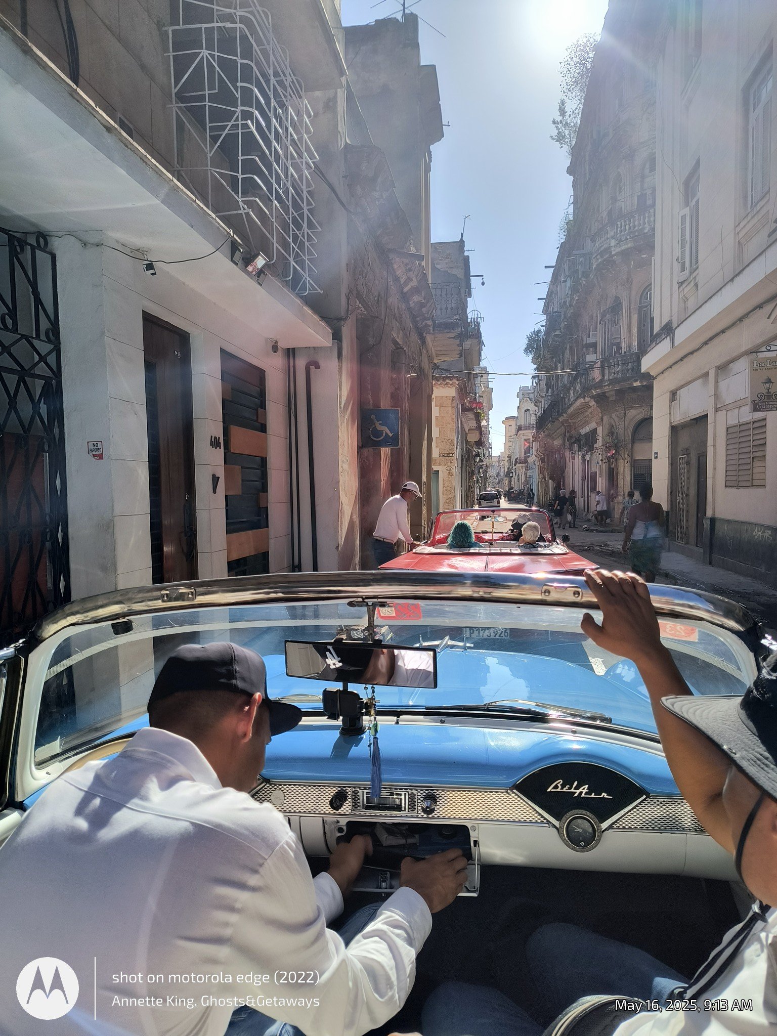 Photo taken from inside a classic blue vintage car showing the dashboard and two people in the front seat, with one man wearing a white shirt and black cap, and another person to the right raising an arm. The street outside has old buildings with bal