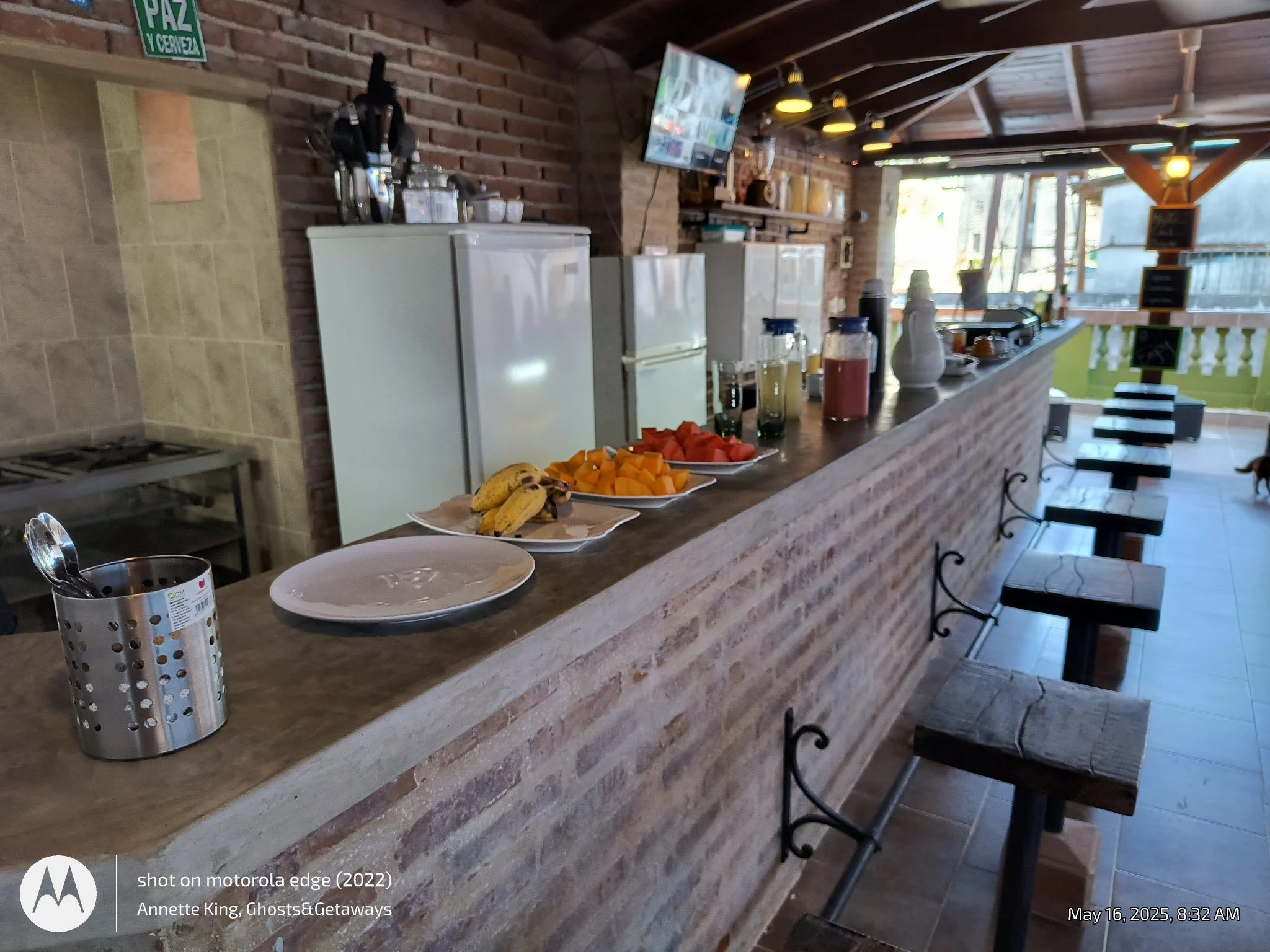 A cozy cafe with a brick and wood interior, featuring a counter with plates of sliced watermelon, bananas, and other fruits, along with condiments and beverages. Bar stools line the front of the counter, and a flat-screen TV is mounted on the brick w