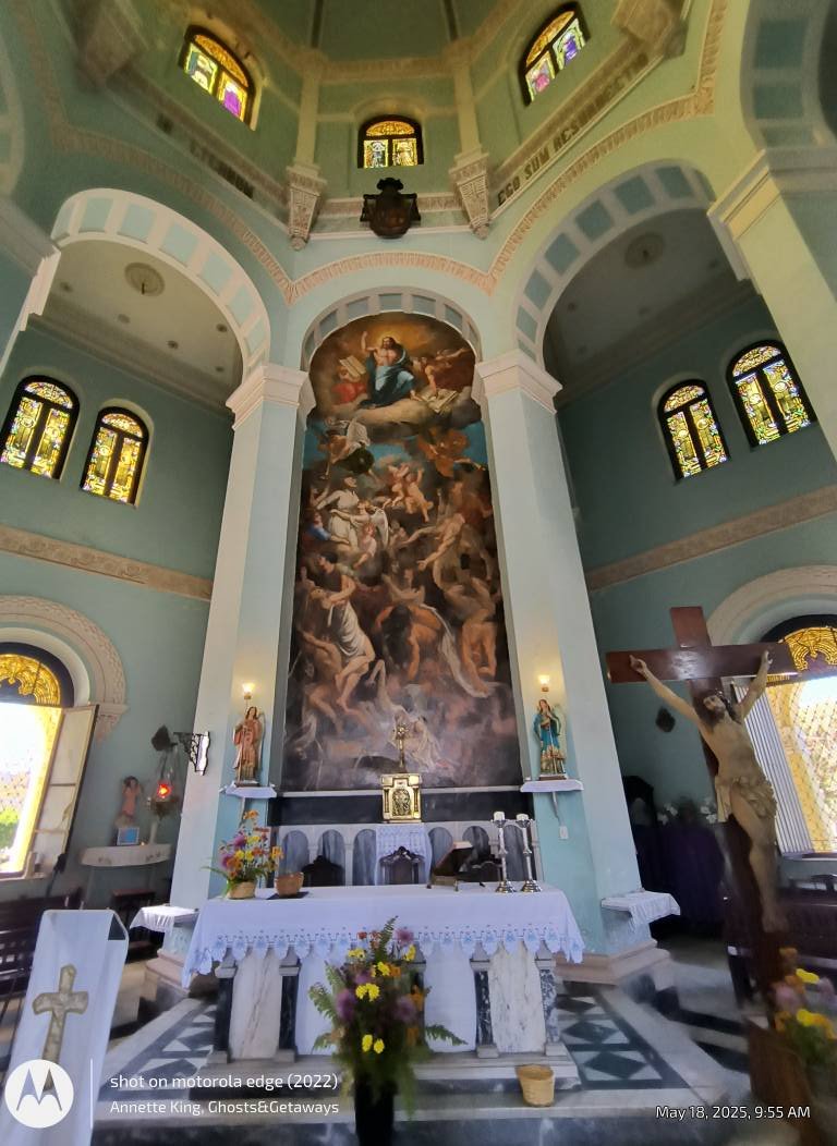 Interior of a church with an ornate altar, a large religious painting depicting heavenly scenes, a crucifix with Jesus on the cross, stained glass windows, and religious statues and items.