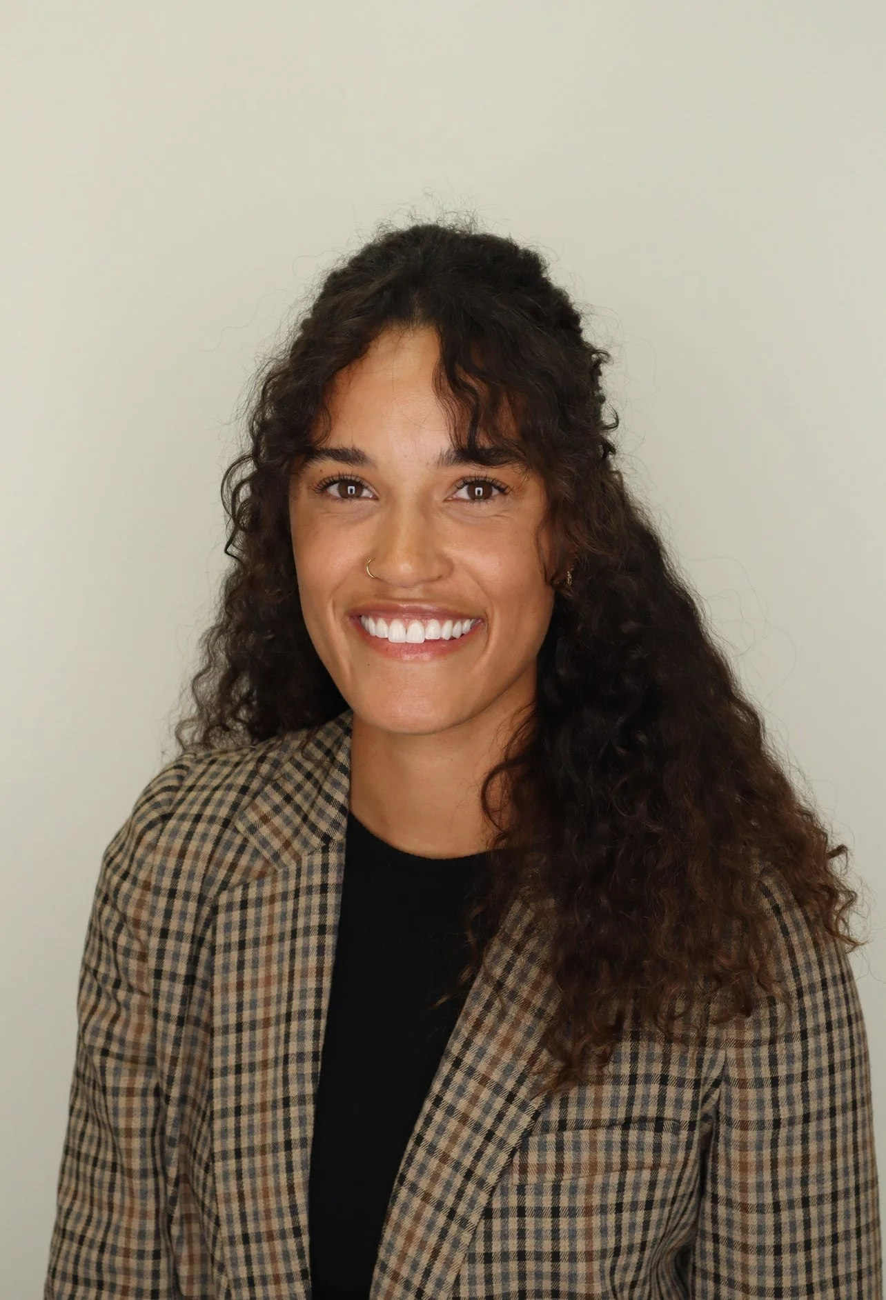 A smiling woman with dark curly hair, wearing a plaid blazer over a black shirt, standing against a plain light-colored background.