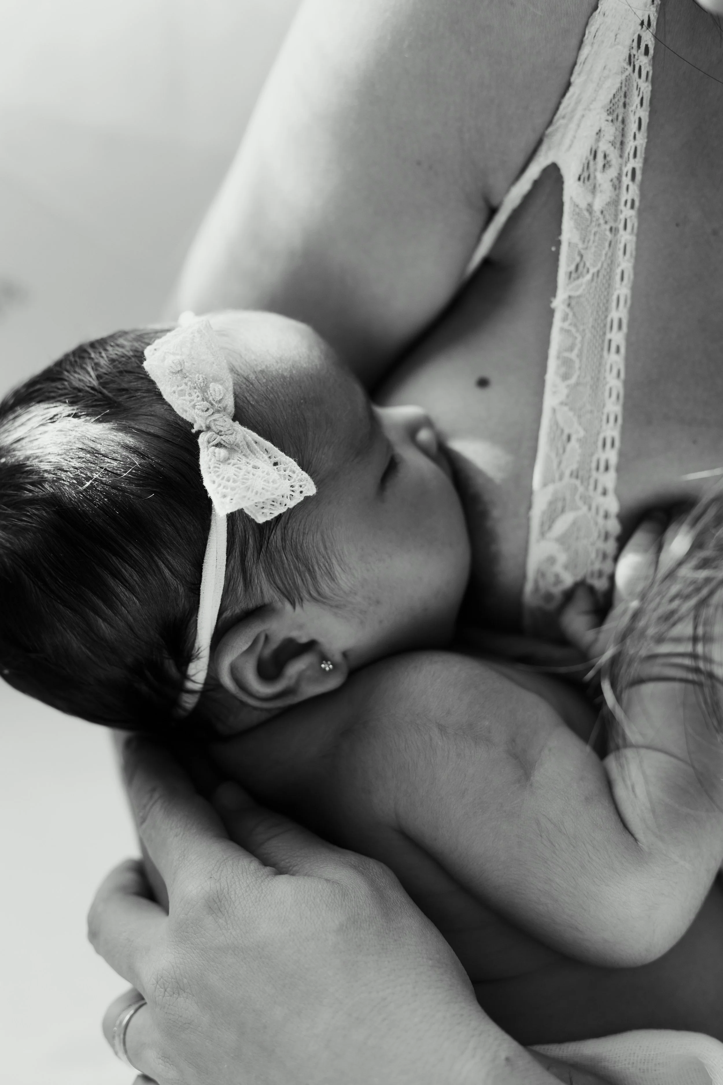 Close-up of a mother breastfeeding her baby, with the baby wearing a lace bow headband and earrings, in black and white.