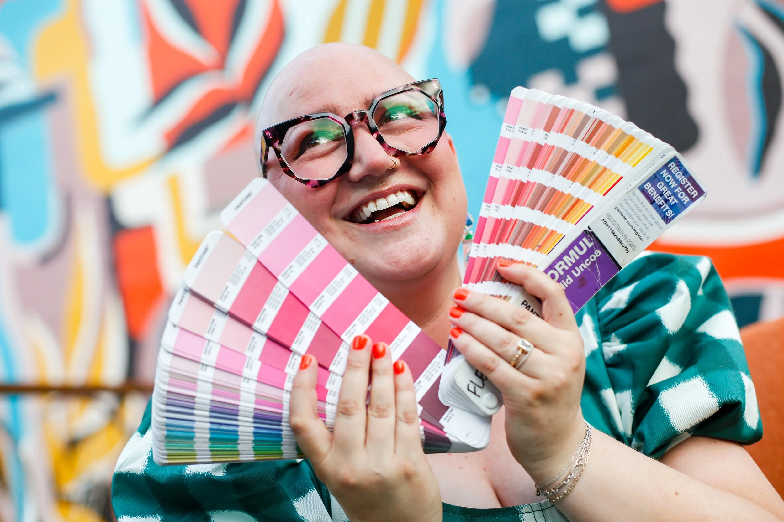 Fleming smiling, holding multiple color swatch books, wearing large glasses, colorful abstract background.