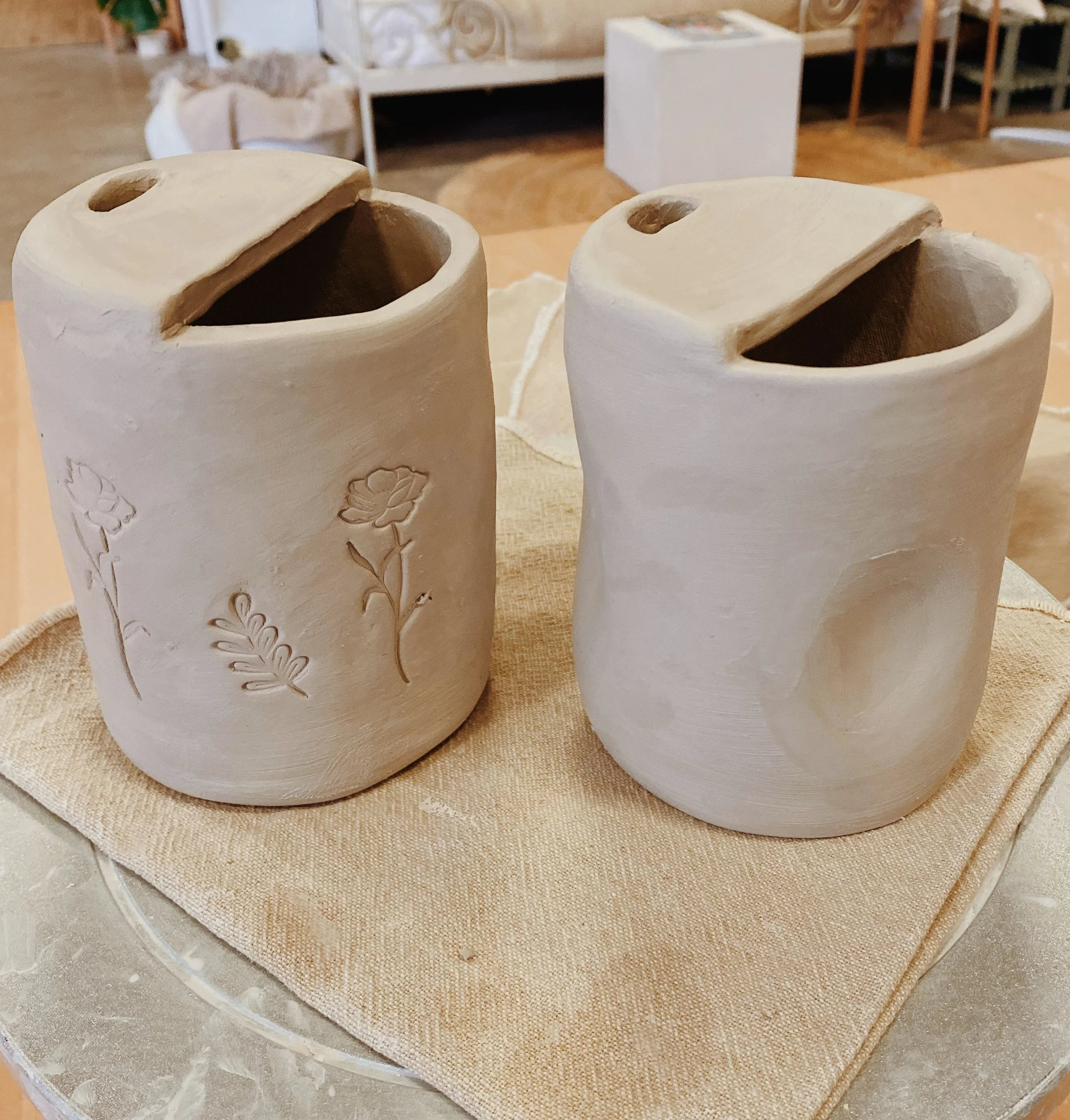 A ceramic vase with a heart-shaped opening and decorative pink designs next to a wooden box with branding, placed on a white surface in front of white beadboard paneling.
