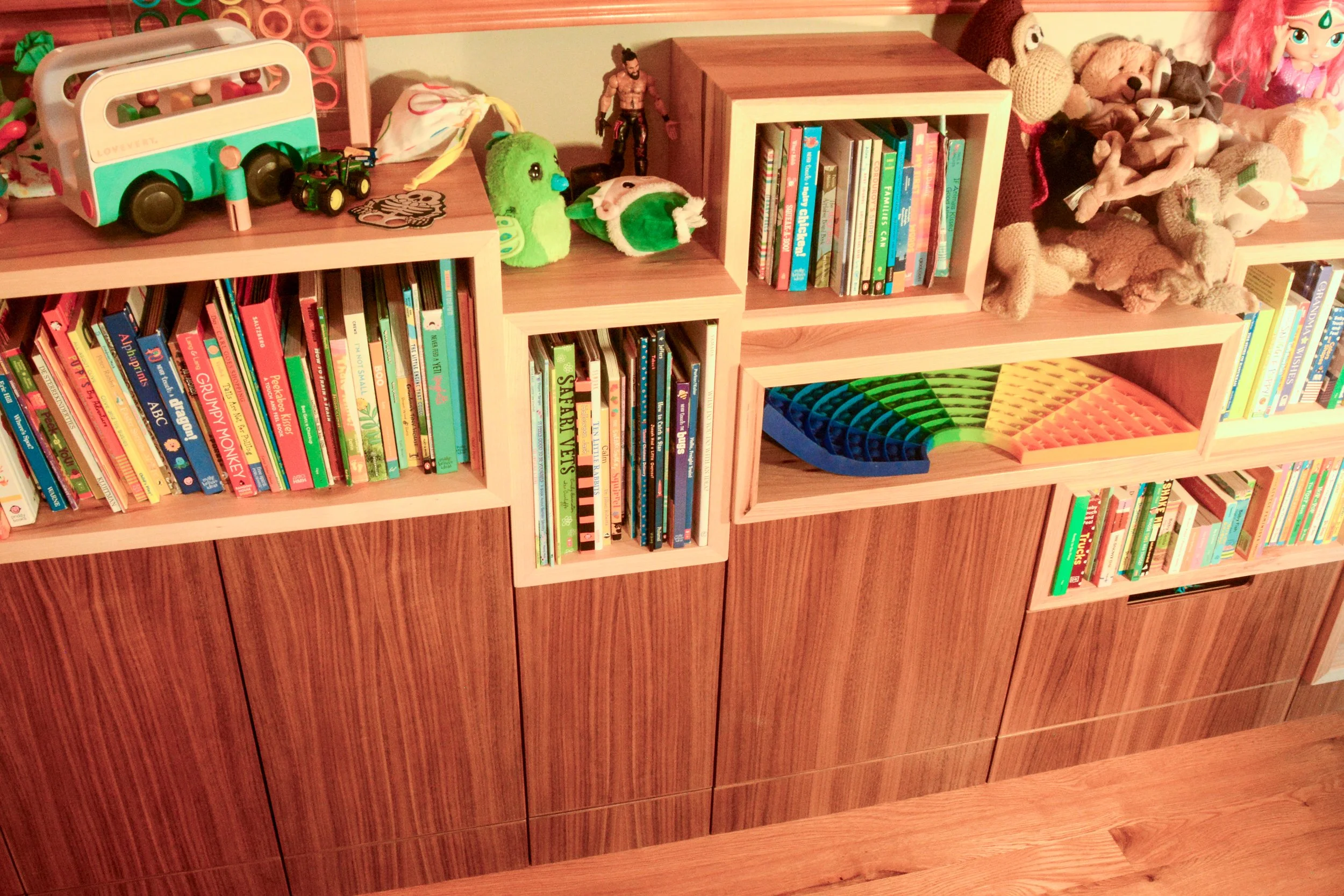 A wooden bookshelf filled with colorful children's books, stuffed animals, toy figures, and a rainbow-shaped wooden puzzle on the shelves.