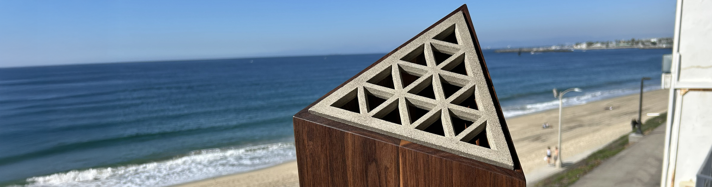 Close-up of a modern architectural element with a triangular concrete and wood structure overlooking a beach and ocean.