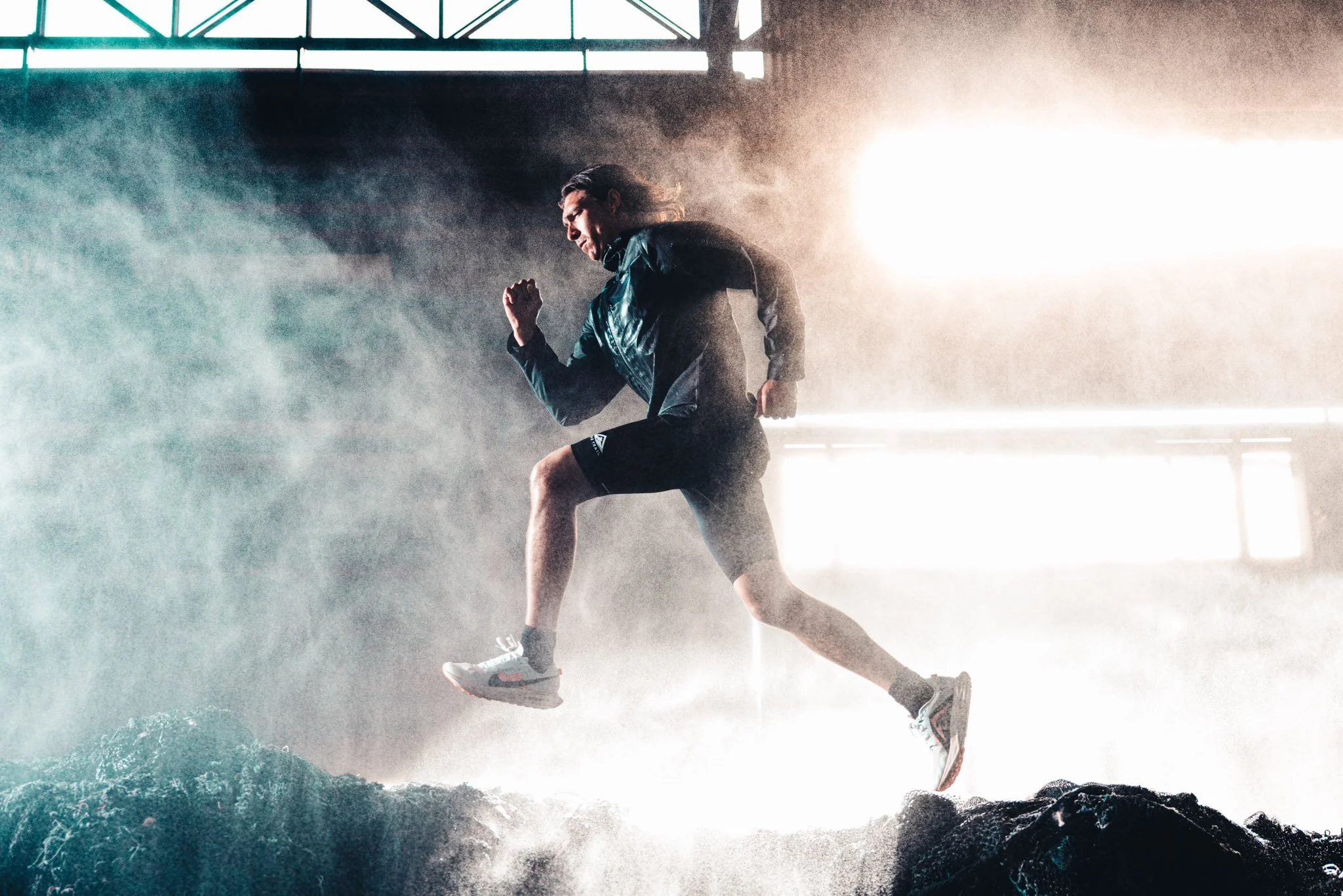 A person is performing a handstand or balancing move on a rocky surface at a misty or smoky environment with bright sunlight in the background.