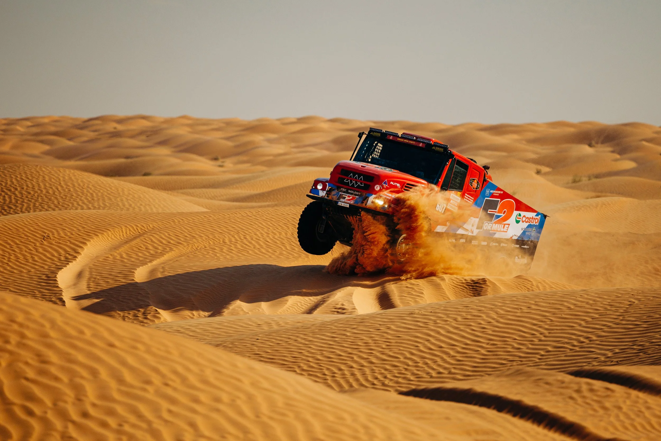 An off-road rally truck speeding through sandy dunes, kicking up sand behind it during daylight.