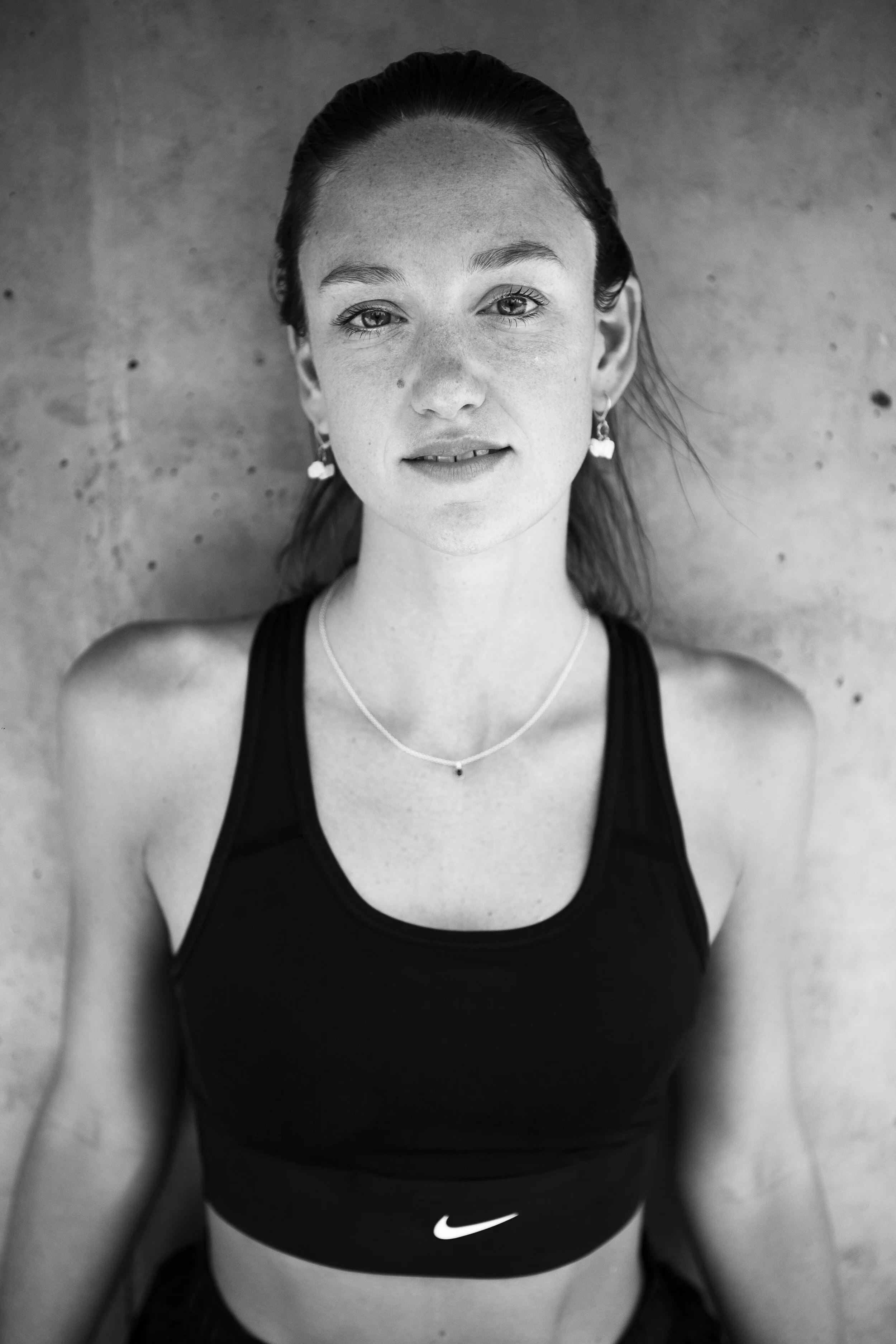 Black and white photo of a young woman with long hair, wearing a Nike sports bra, earrings, and a necklace, looking directly at the camera with a slight smile, standing against a concrete wall.