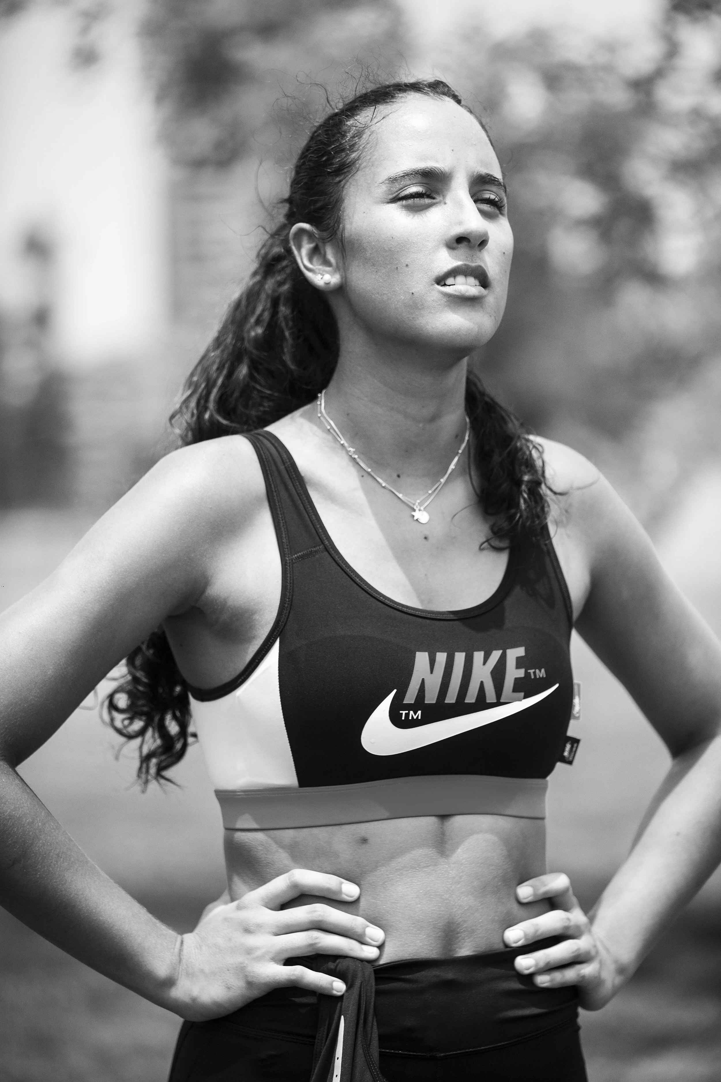 Black-and-white photo of a female athlete in athletic wear, standing outdoors with her hands on her hips, looking off into the distance with a determined expression.