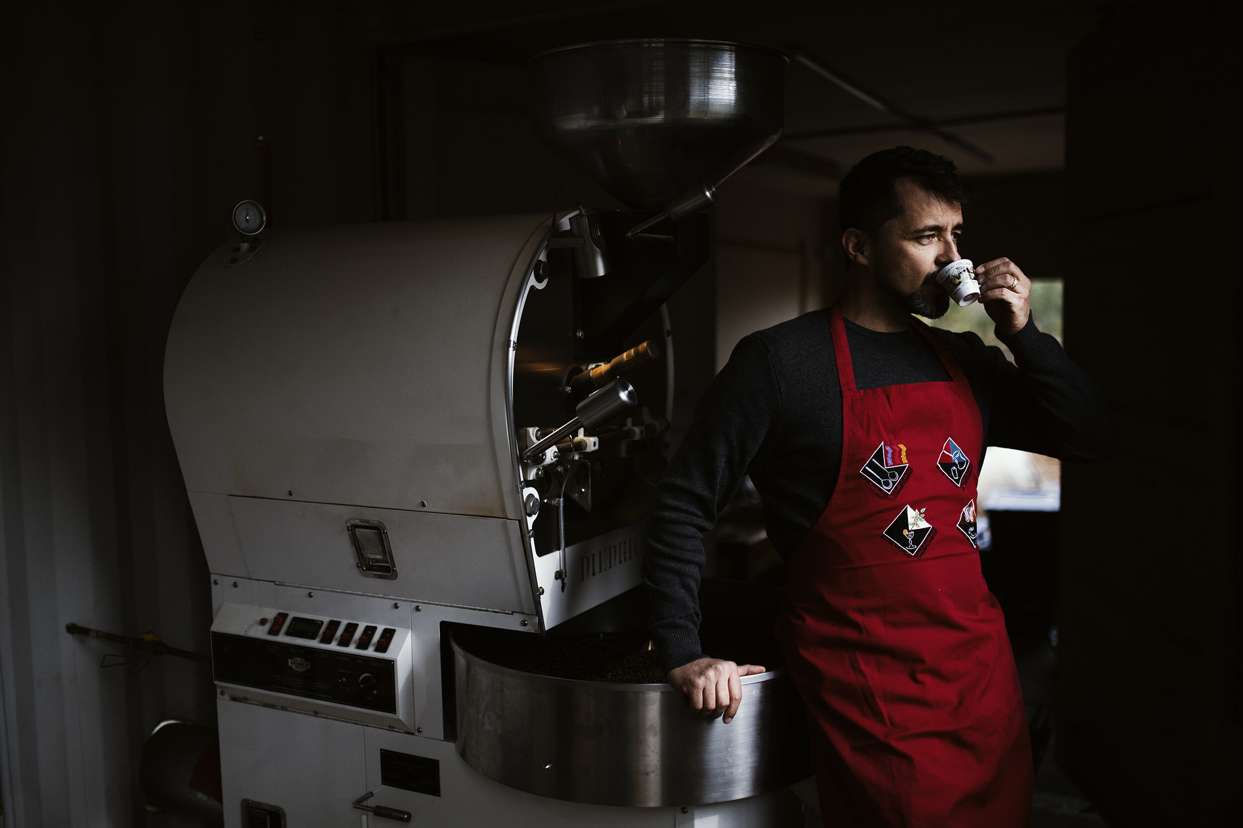 Man wearing a red apron and drinking coffee next to a large coffee roasting machine.