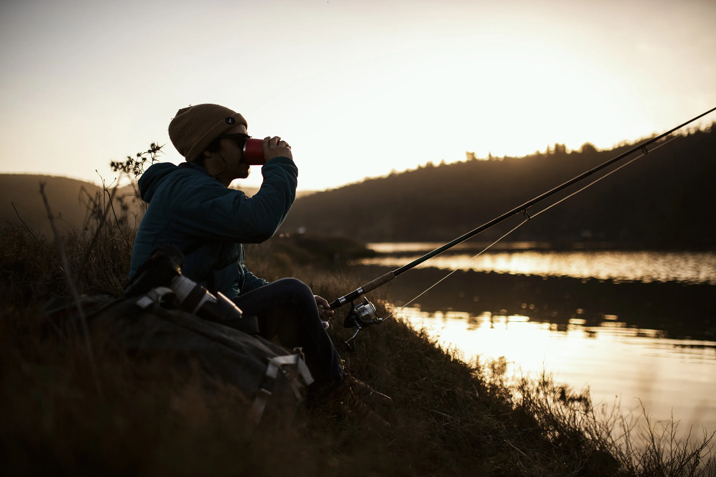 Person sitting by a riverbank at sunset, fishing and drinking from a cup.
