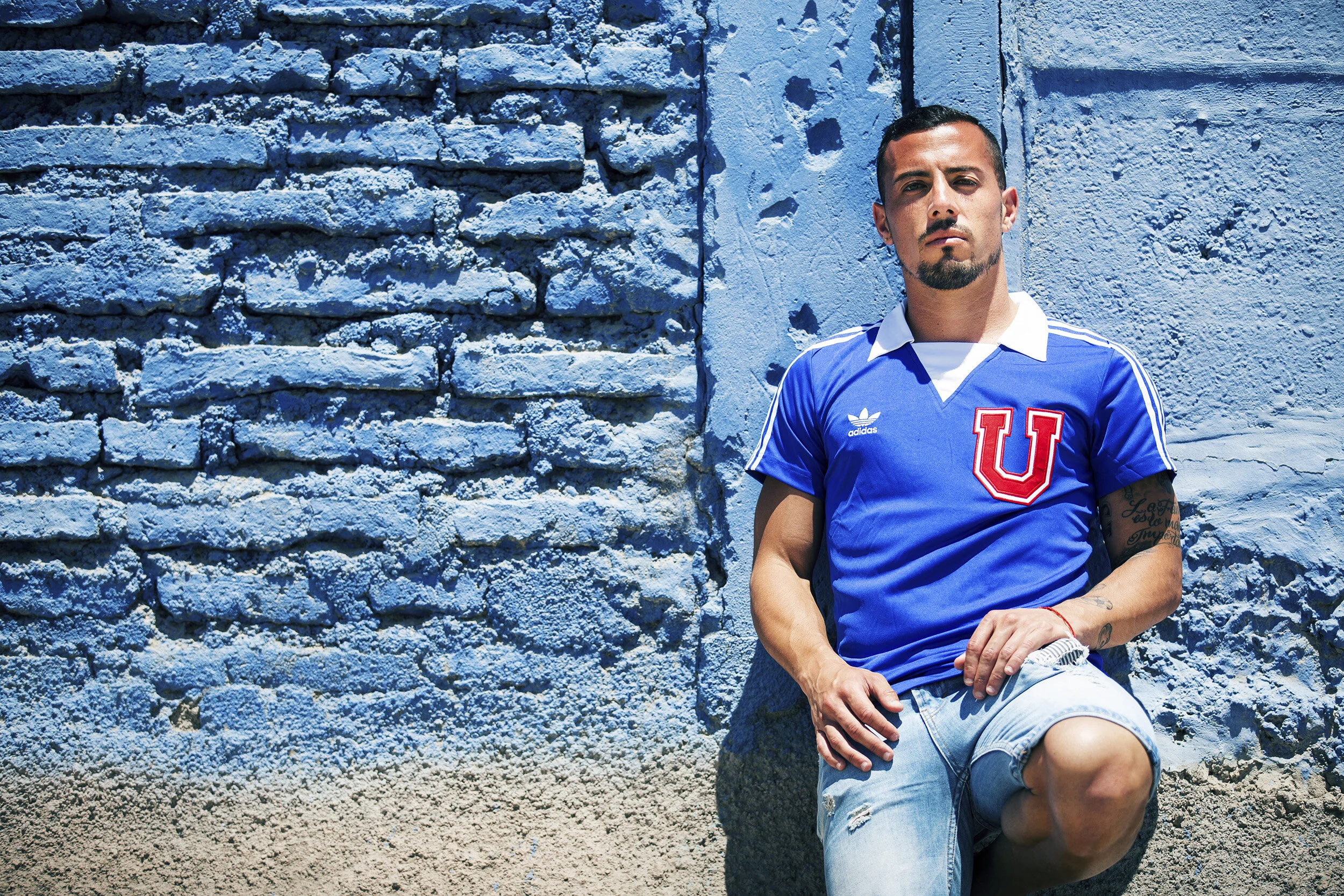 A man kneeling against a textured blue wall, wearing a blue soccer jersey with a large red 'U' on it and ripped denim shorts.