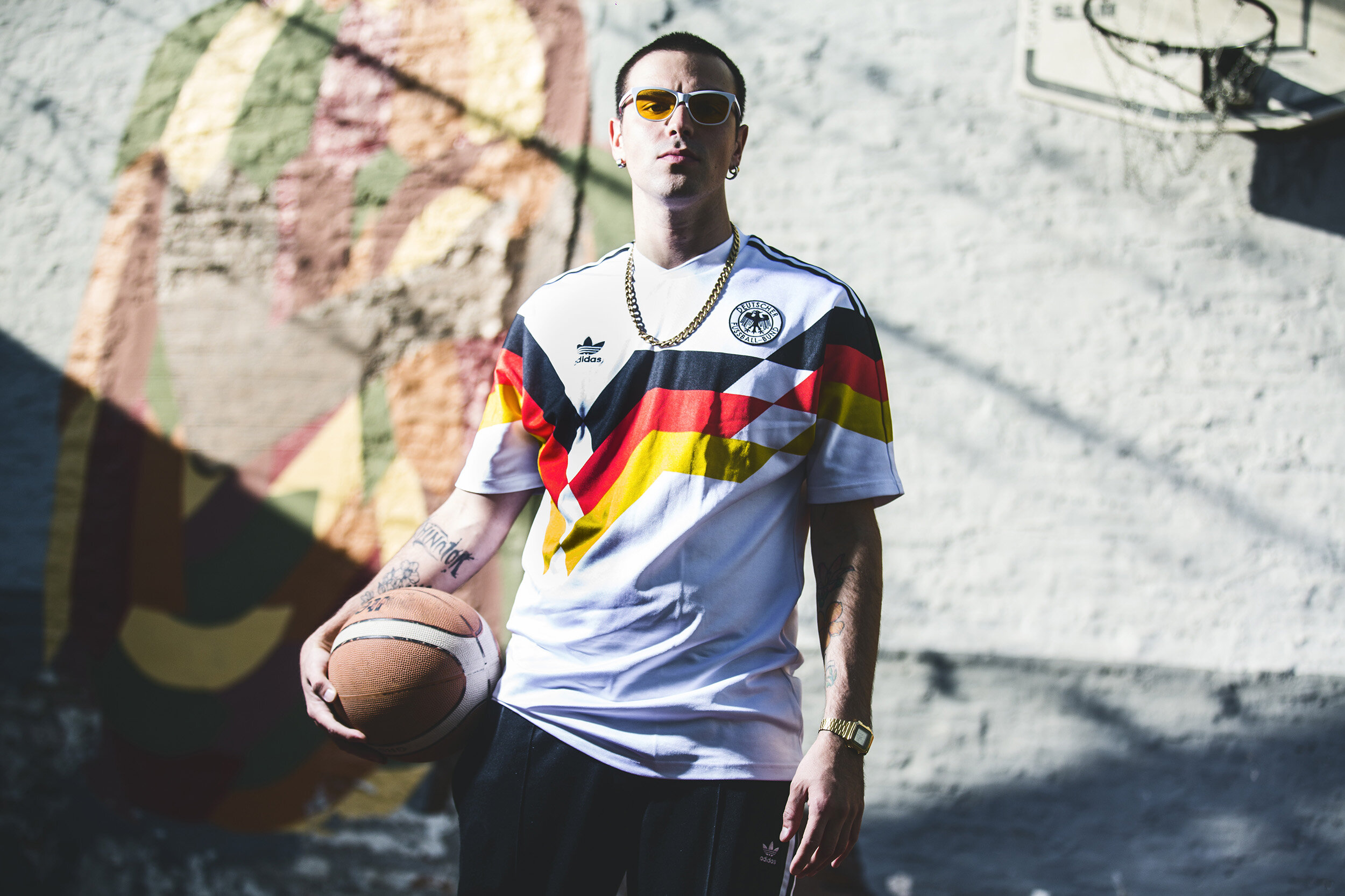 A young man with sunglasses holding a basketball, standing outdoors against a brick wall with a colorful mural, wearing a Germany soccer jersey, gold chain, and a watch.