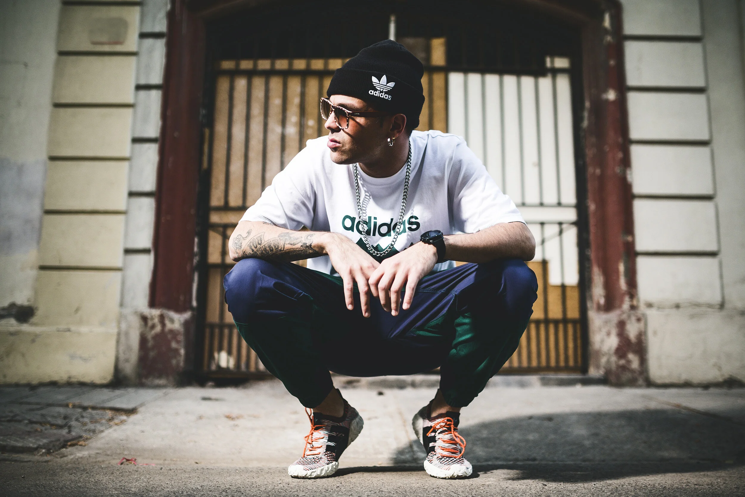 A man squatting outdoors in front of a gate and wall, wearing a black beanie with Adidas logo, sunglasses, a white Adidas t-shirt, dark pants, and athletic shoes, with tattoos on his arm and wearing accessories.