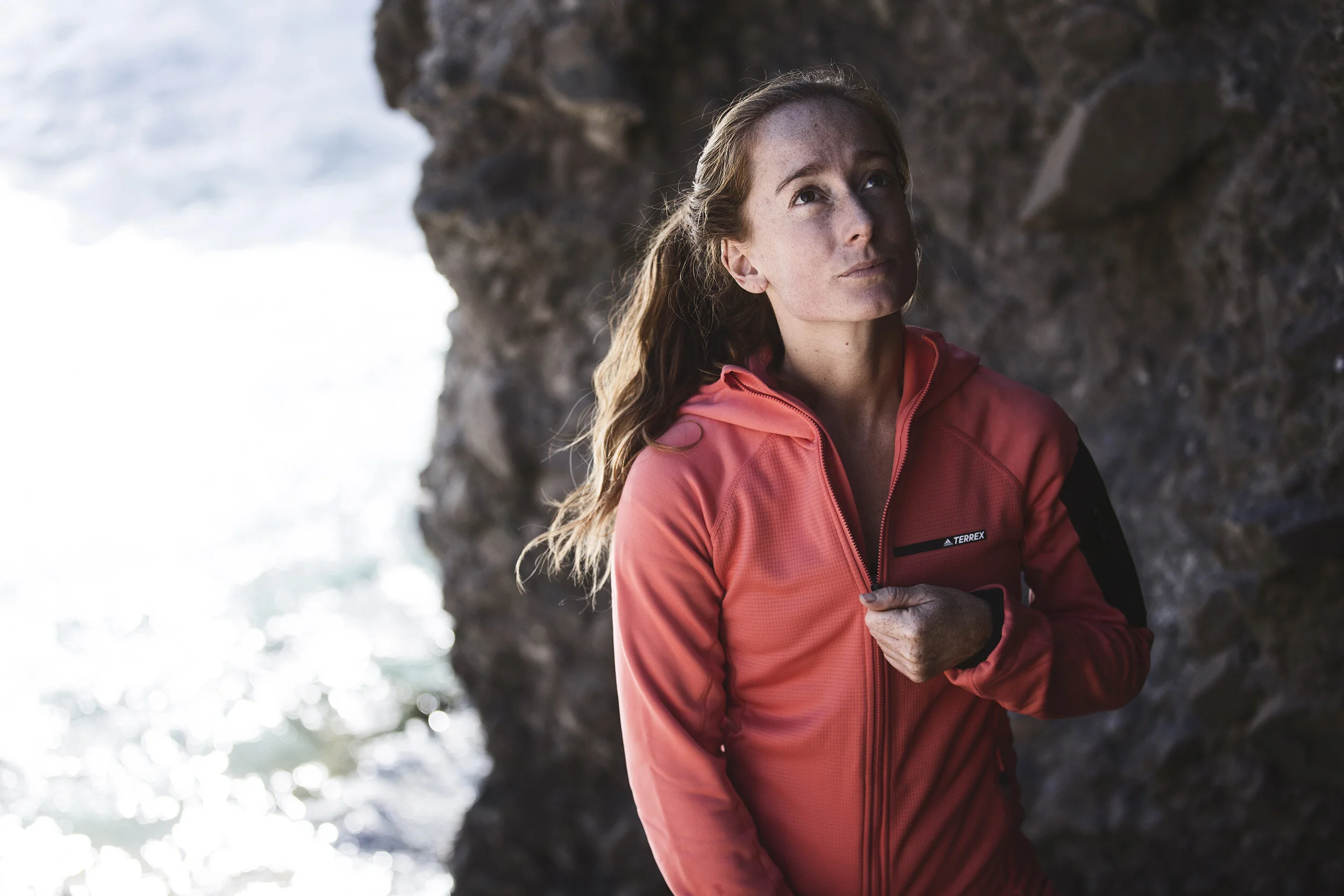 A woman with long hair wearing a pink jacket outdoors near a rocky cliff with water in the background.